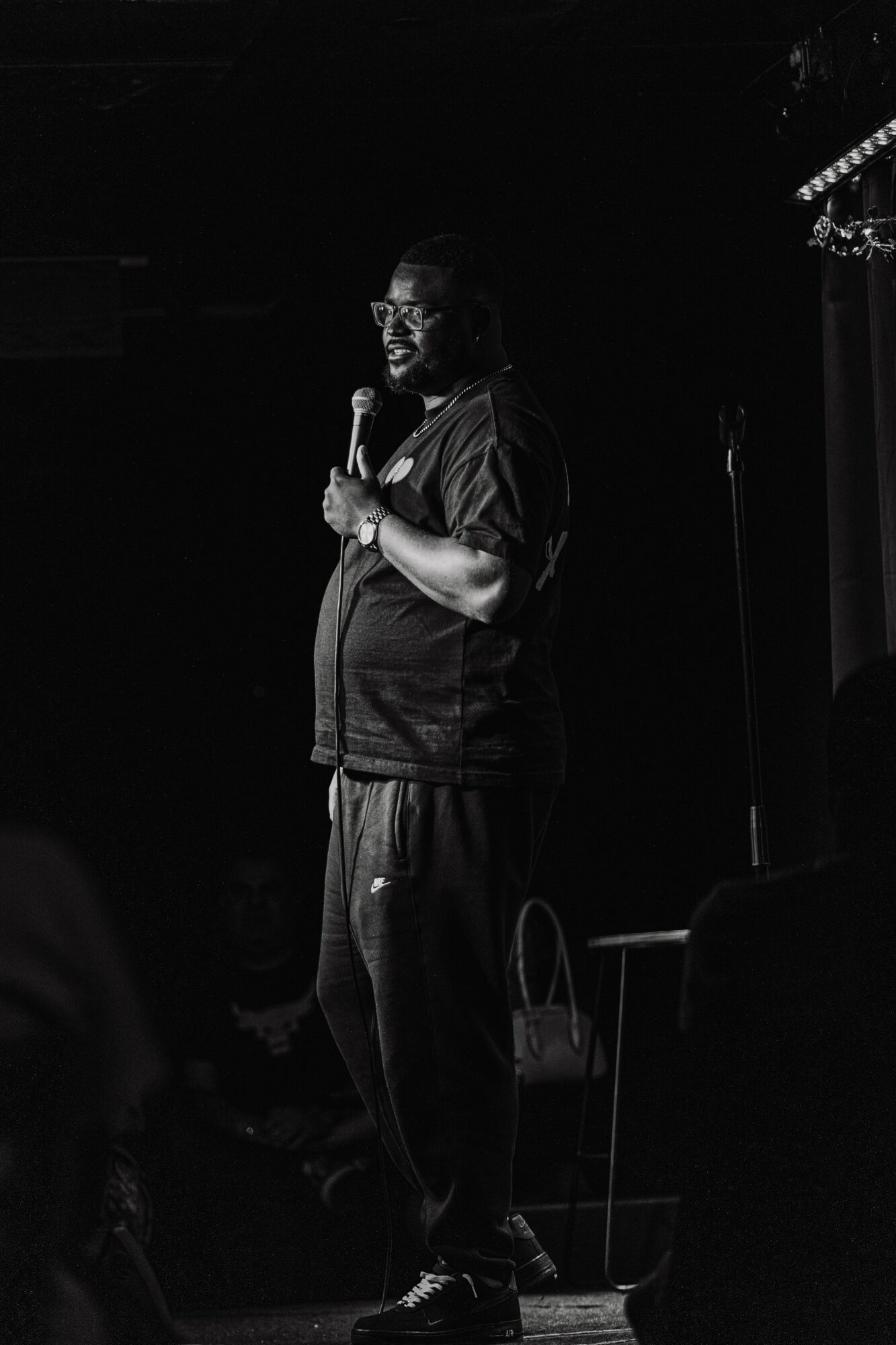 Person holding a microphone on stage, smiling, with a dark background and stage lighting, standing near a chair.
