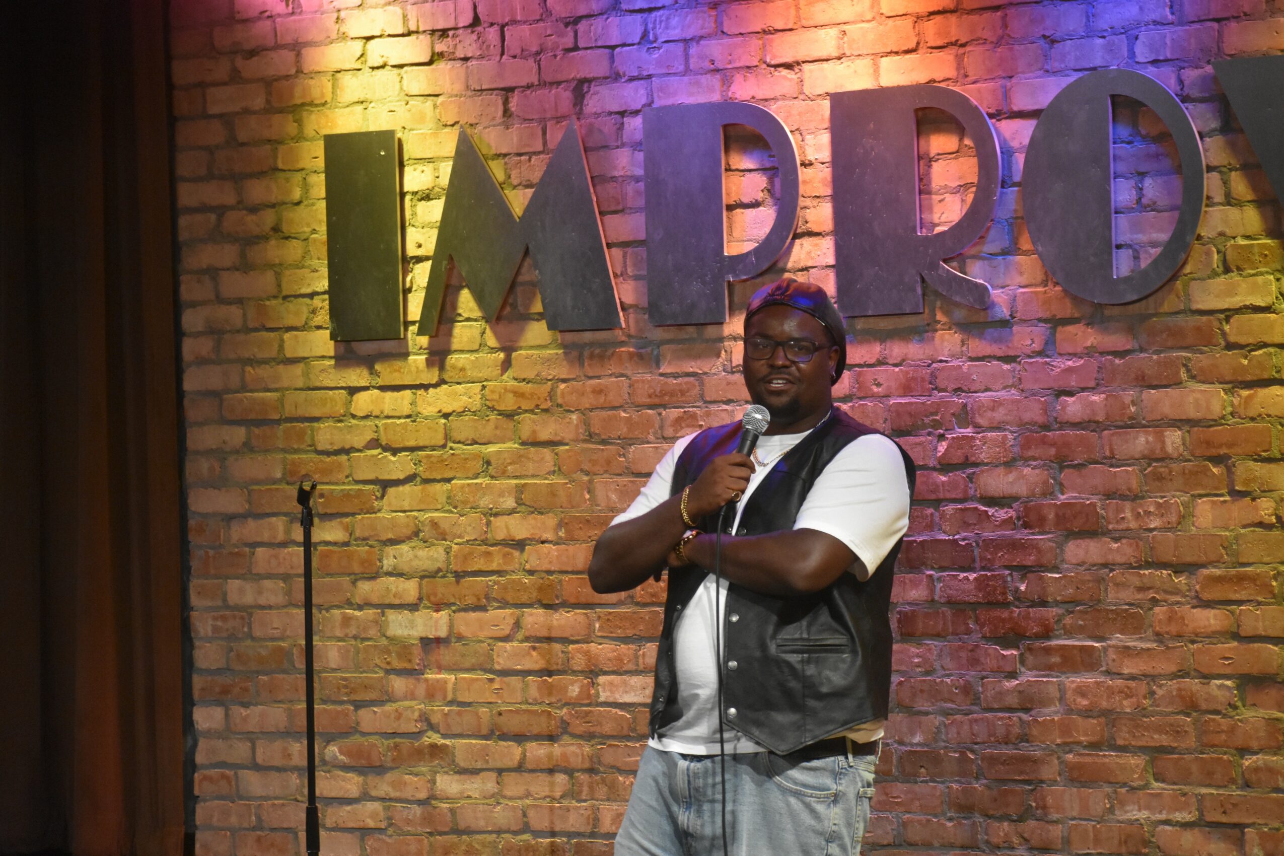 Person standing on stage with a microphone in front of a brick wall with large letters spelling 'IMPRO'.