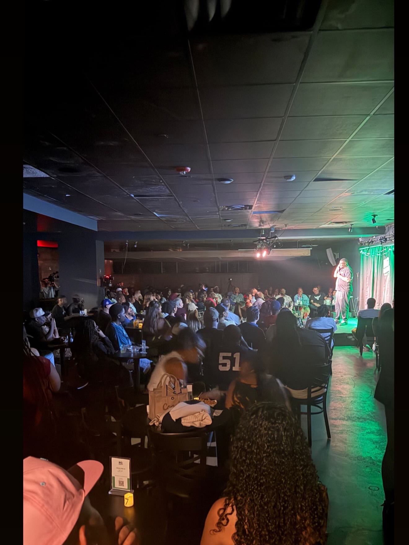 Audience watching a performer on stage in a dimly lit venue with green lighting, seated at tables, some with drinks.