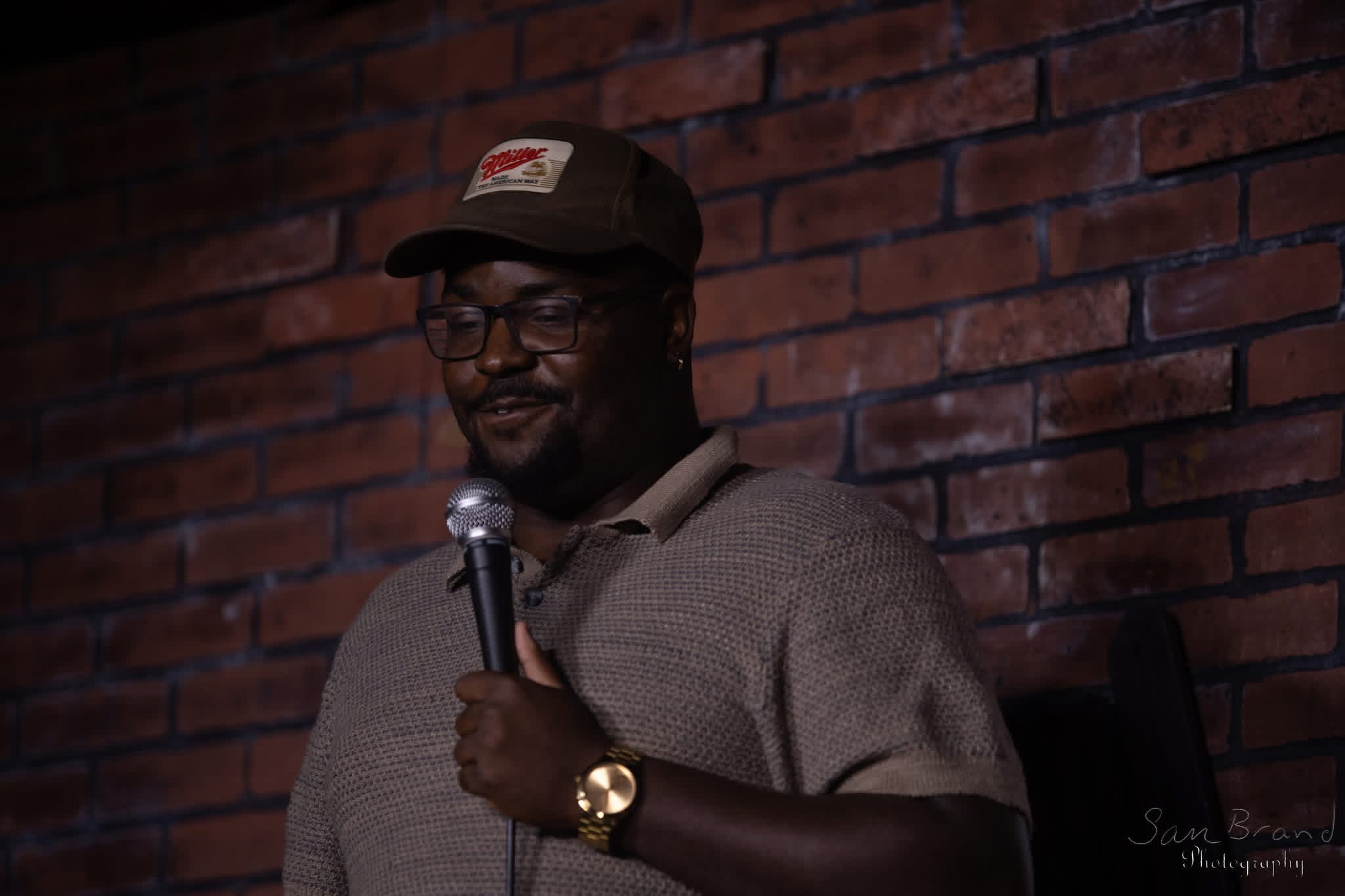 Man wearing glasses, a cap, and a watch, holding a microphone, standing in front of a brick wall.