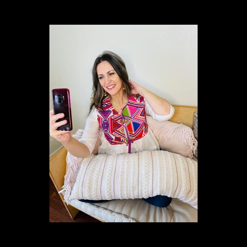 Woman taking a selfie with a smartphone, smiling, sitting on a bed with pillows, against a plain wall.