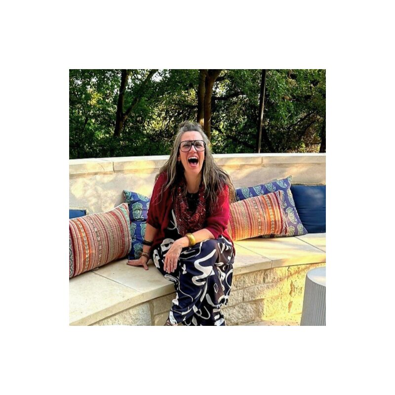 Woman with glasses and long hair sitting on a stone bench outdoors, smiling, surrounded by colorful pillows, trees in background.