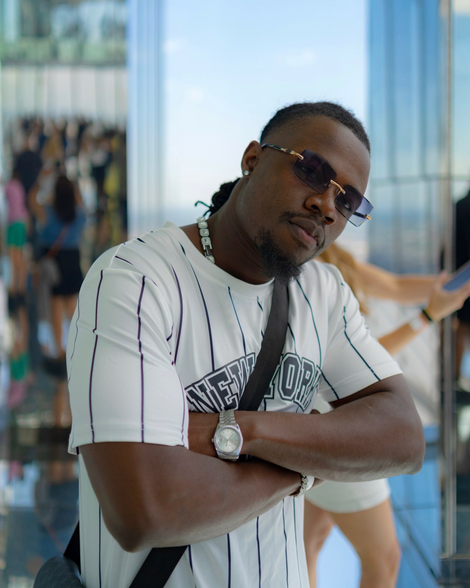 Man with sunglasses and a white striped shirt crossing arms indoors, with people and windows in background.