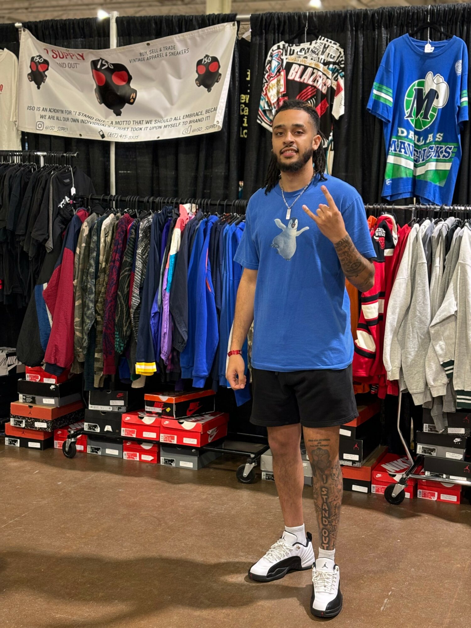 Young man with dreadlocks and a beard stands in a clothing store, wearing a blue t-shirt, black shorts, and white sneakers, making a hand gesture.