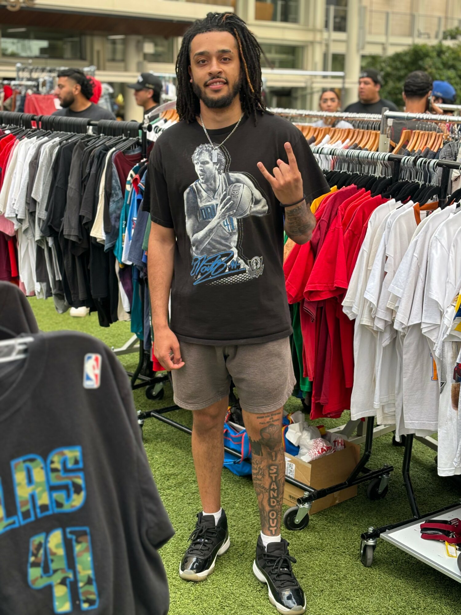 Young man with dreadlocks and a beard standing outdoors among clothing racks, making a hand gesture, wearing a graphic t-shirt and shorts.
