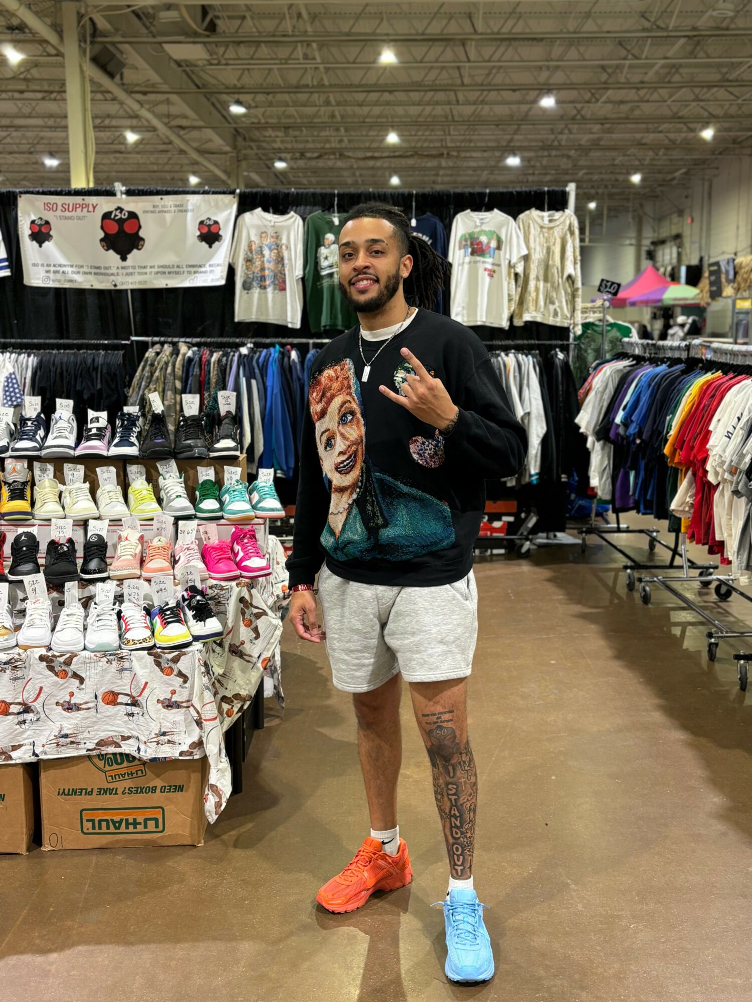 Young man with a beard and tattoos, wearing a black jacket, white shorts, and colorful sneakers, standing in a clothing store.