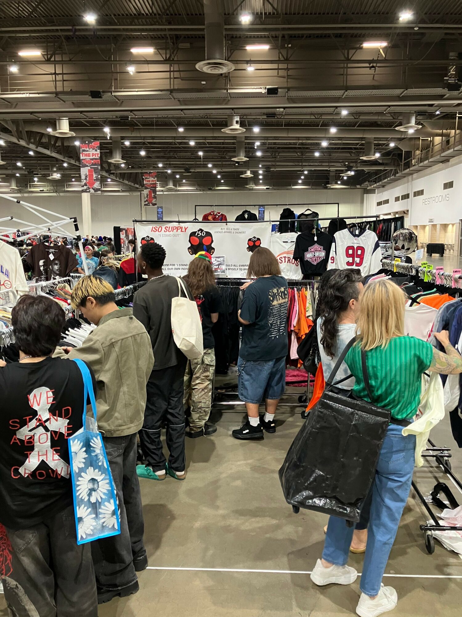 People standing in line at an indoor event, some holding bags, with bright ceiling lights and a busy atmosphere.