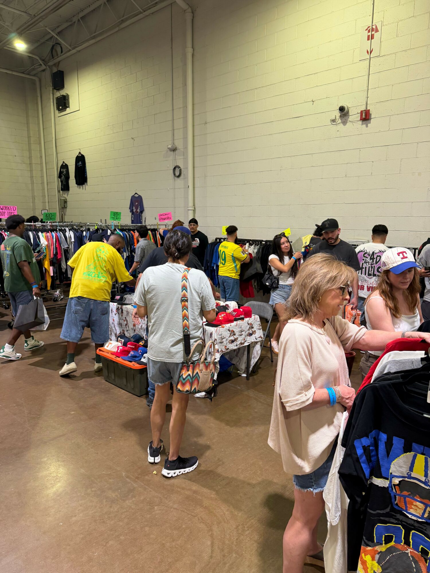 People gather around tables in a large indoor space, possibly a community event or market, with high ceilings and bright lighting.