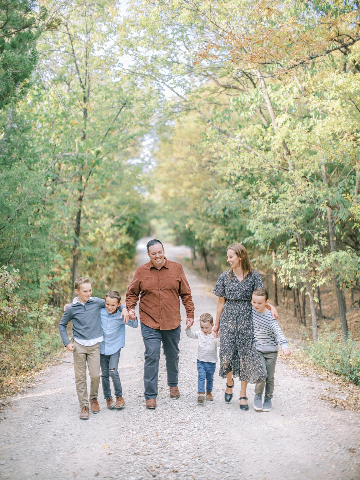 Family of six walking on a dirt path surrounded by trees with green and yellow leaves, holding hands and talking.