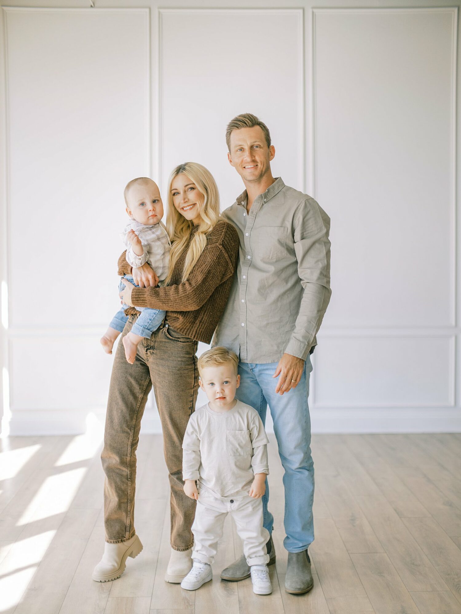 Family of four standing in a bright room with white walls, smiling, with sunlight streaming in, including a woman, man, and two children.