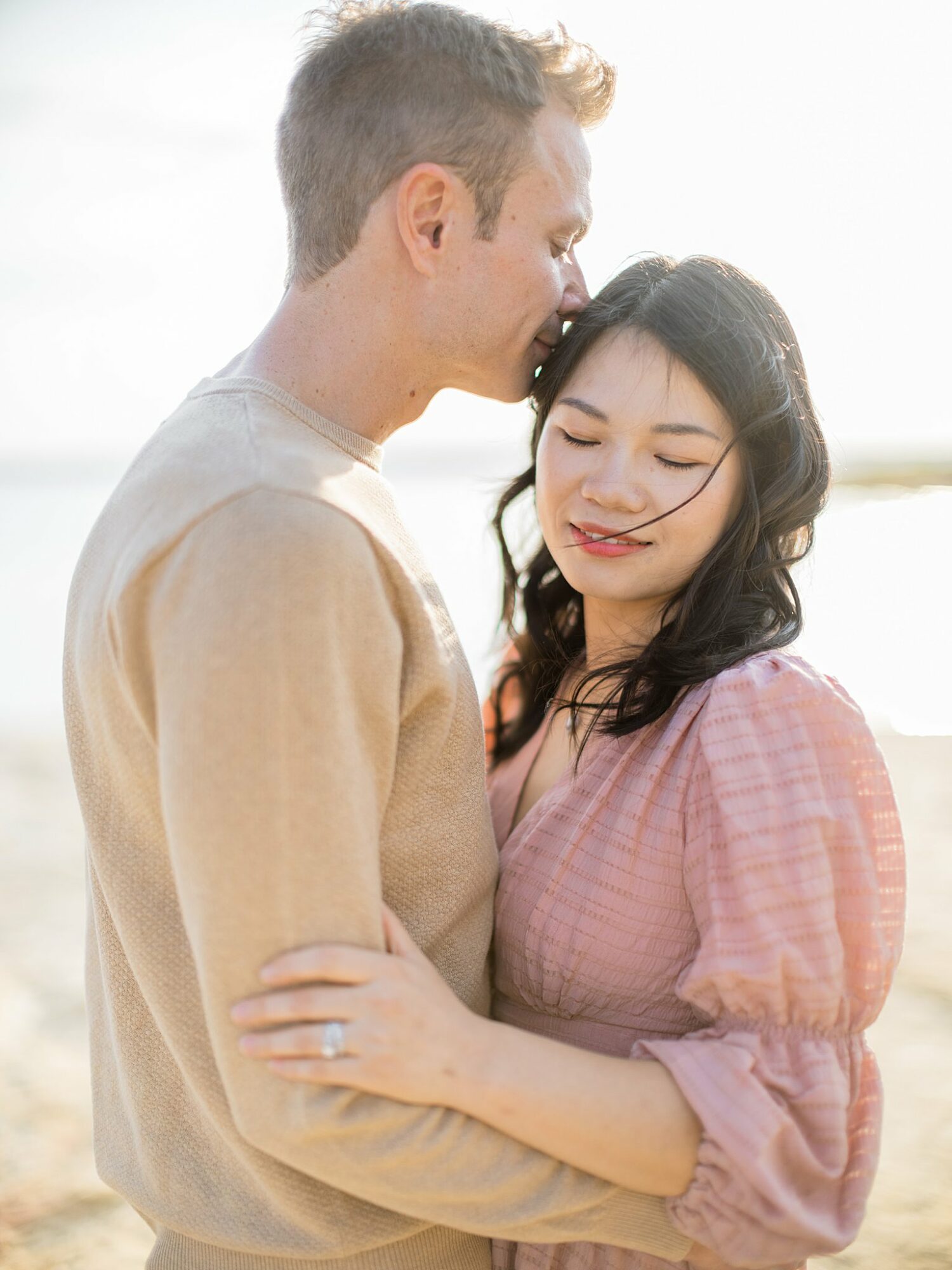 A man and woman embrace, the man kisses the woman's forehead outdoors with bright light behind them.