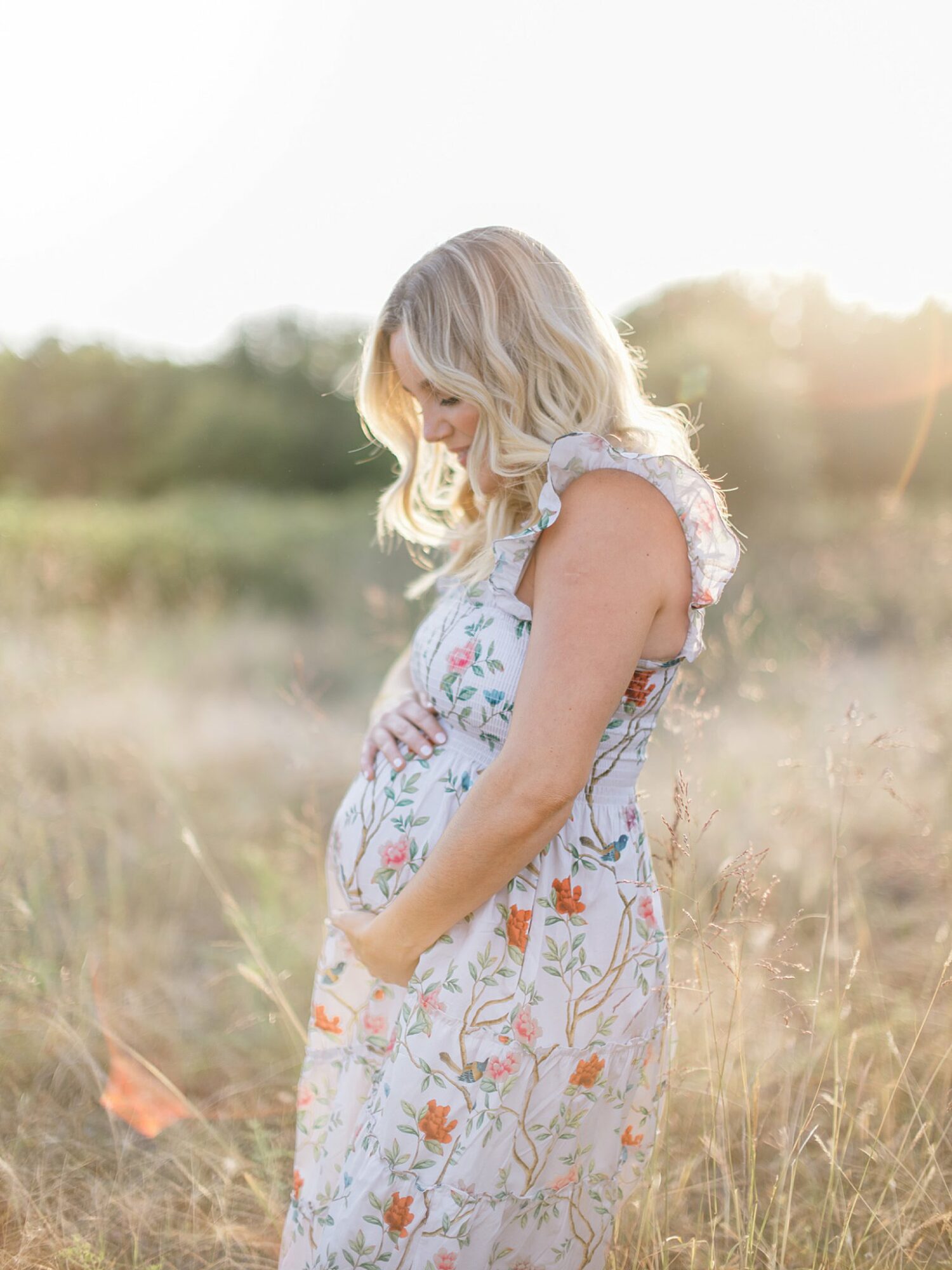 Young woman with blonde hair in a floral dress standing in a field with sunlight behind her.
