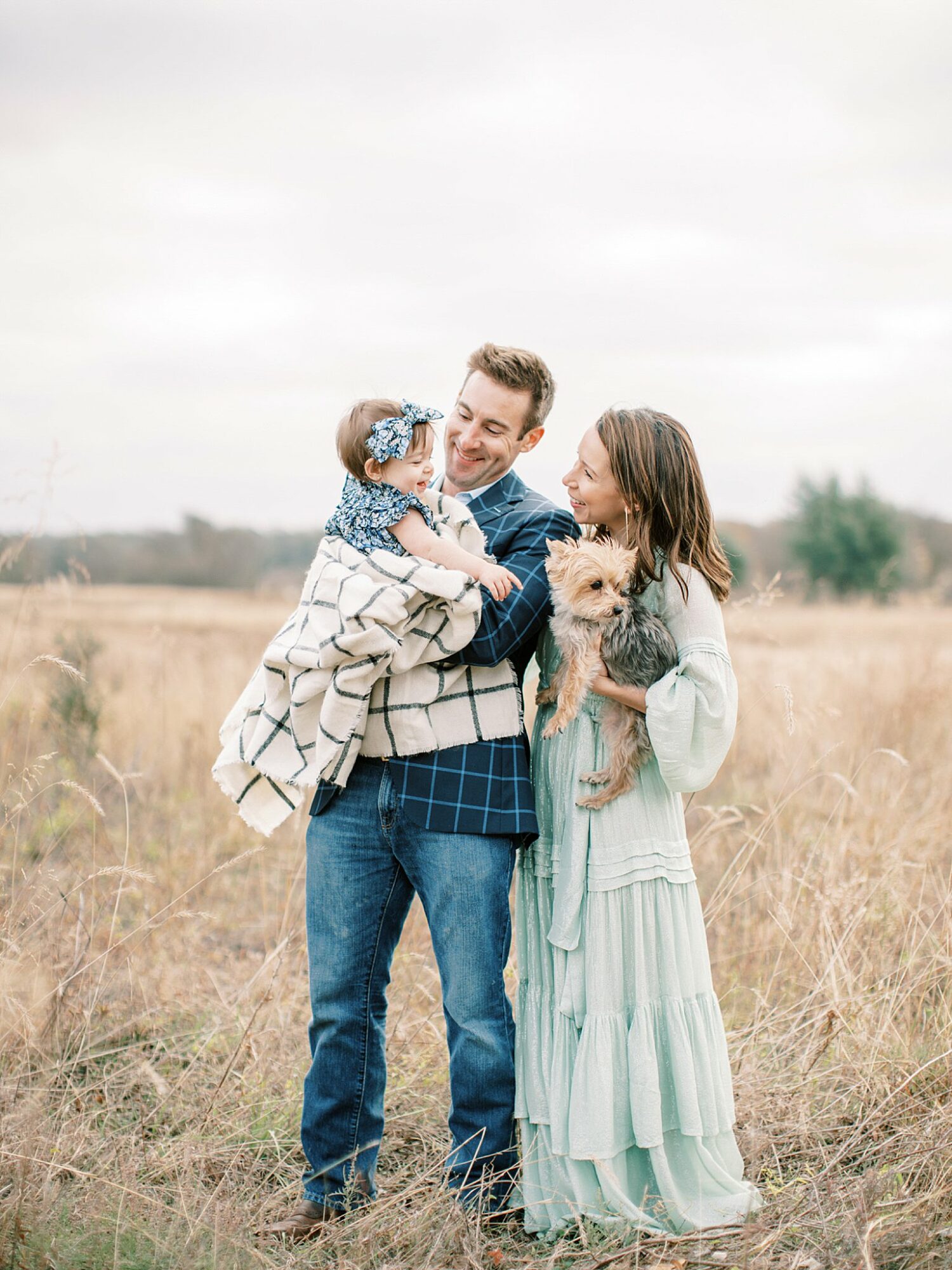 Family of three with a small dog standing in a field, smiling and interacting outdoors on a cloudy day.