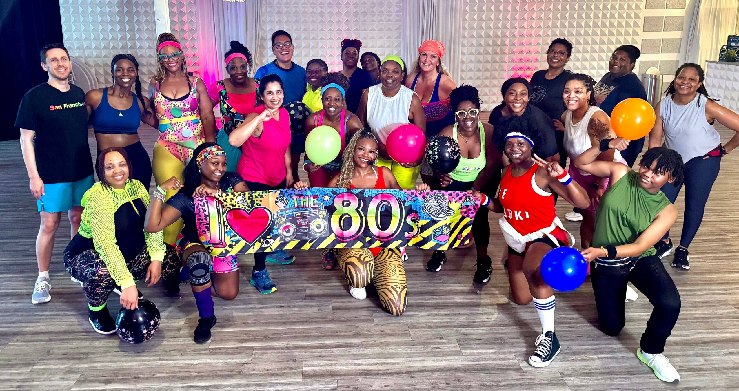 Group of people celebrating an 80s themed party with balloons and colorful clothing, holding a banner, in a room with a patterned wall.