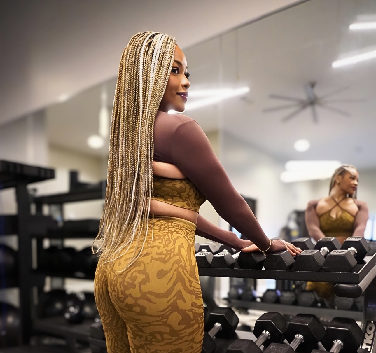 Woman with long braided hair in a gym, looking at dumbbells, wearing a yellow patterned outfit, smiling, mirror reflection visible.