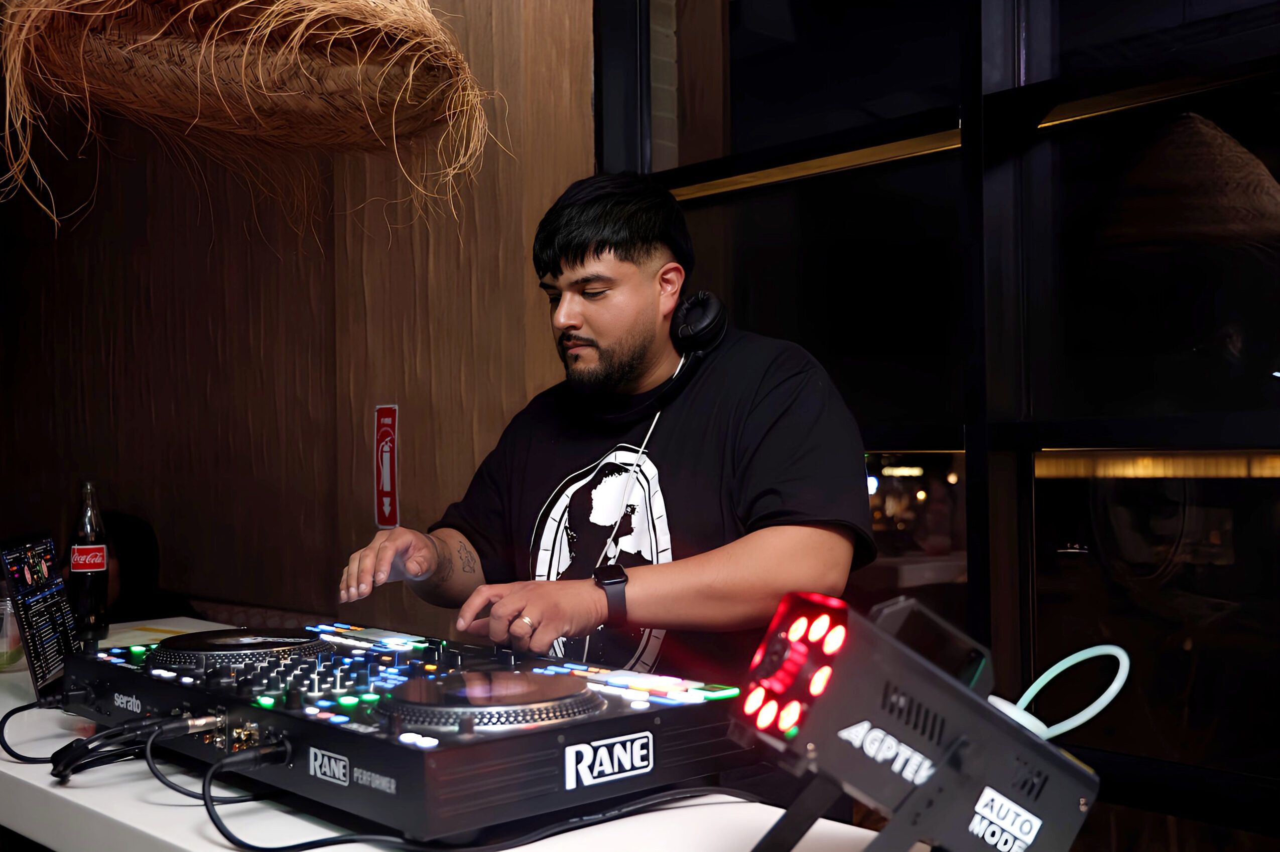 DJ with headphones operating a mixing console in a dimly lit indoor space, with a window showing darkness outside.