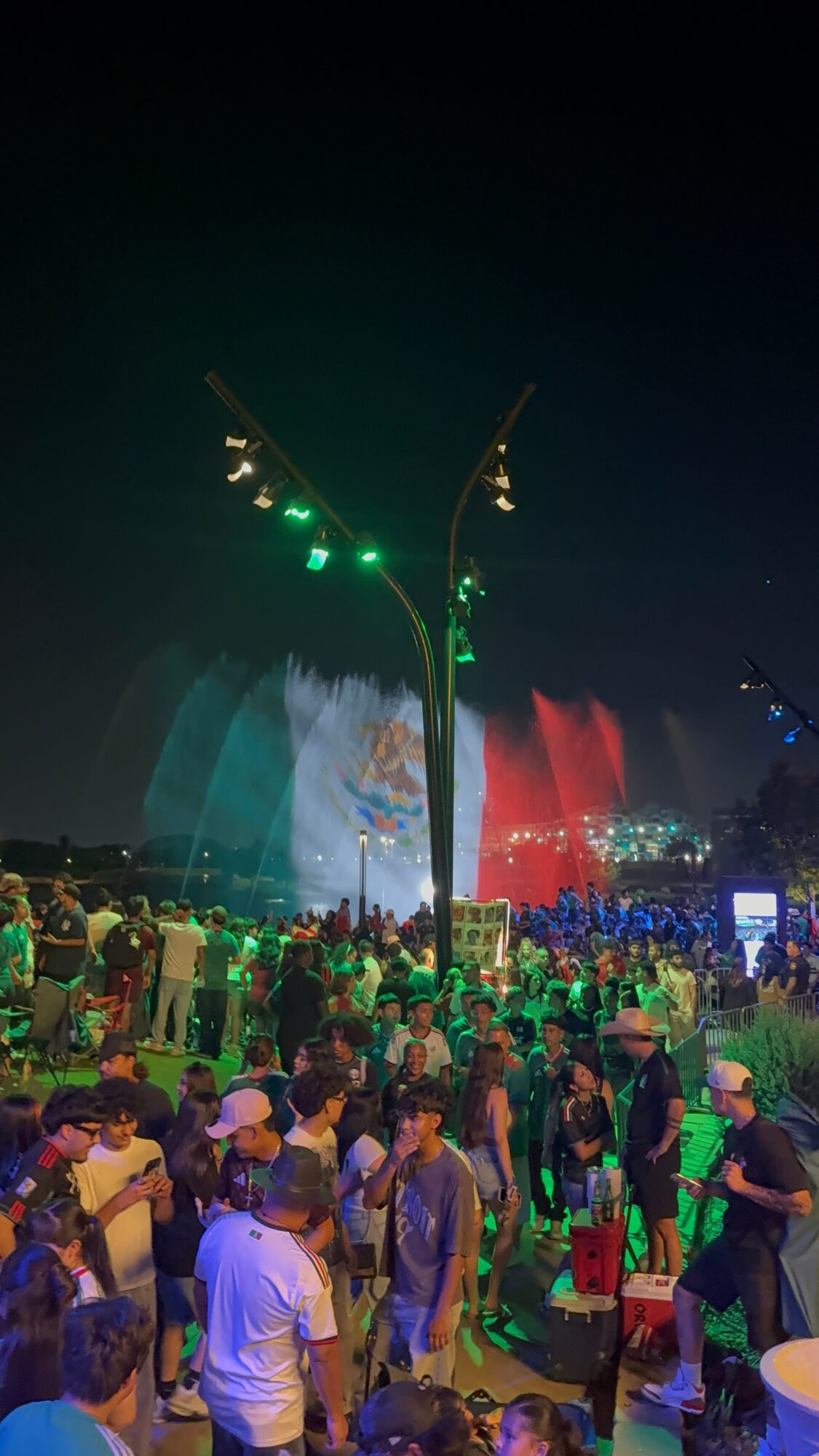 Crowd watching a colorful fountain display at night with lights and water jets, illuminated by green and red lights.