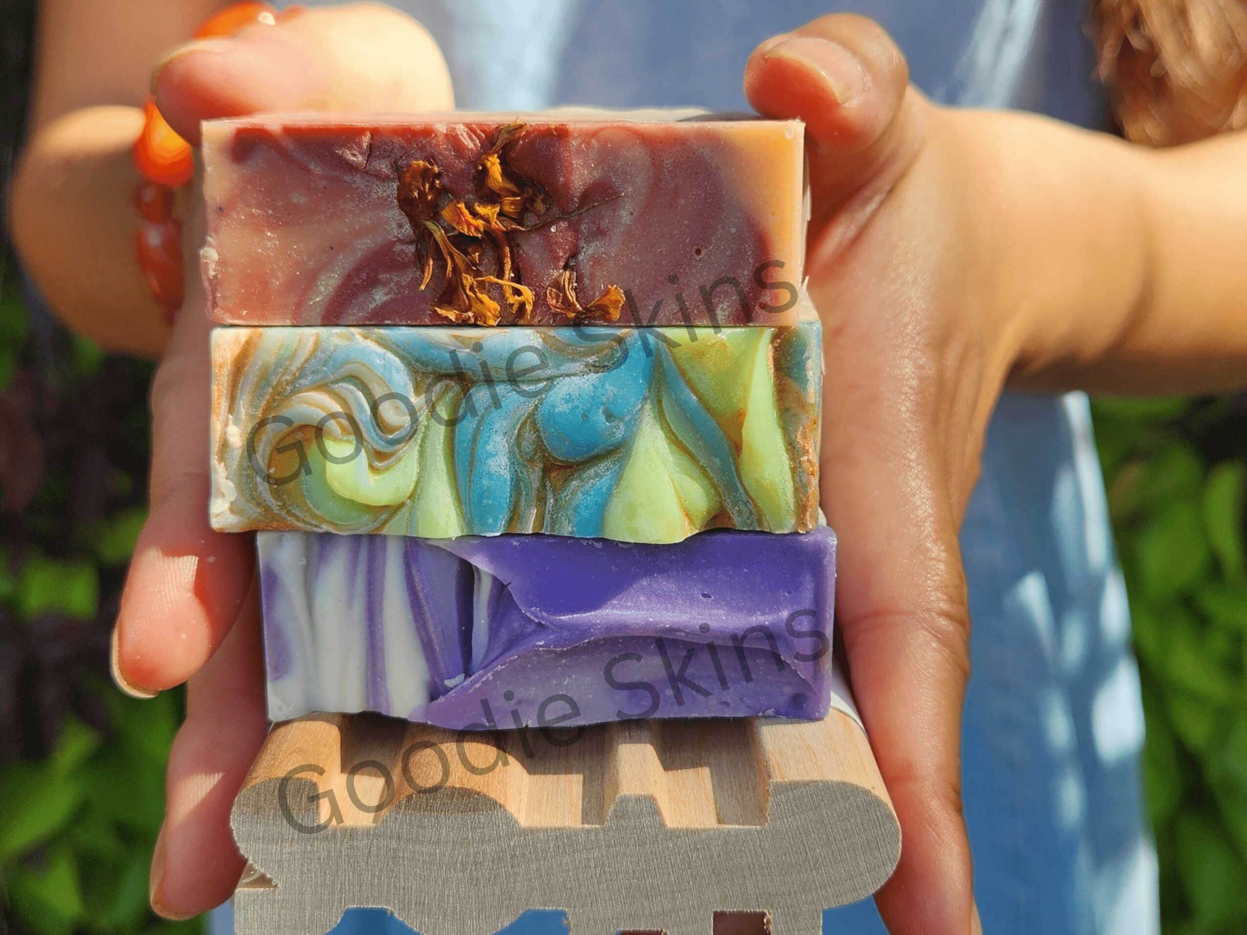 Person holding three colorful soap bars outdoors, with a blurred background of greenery.