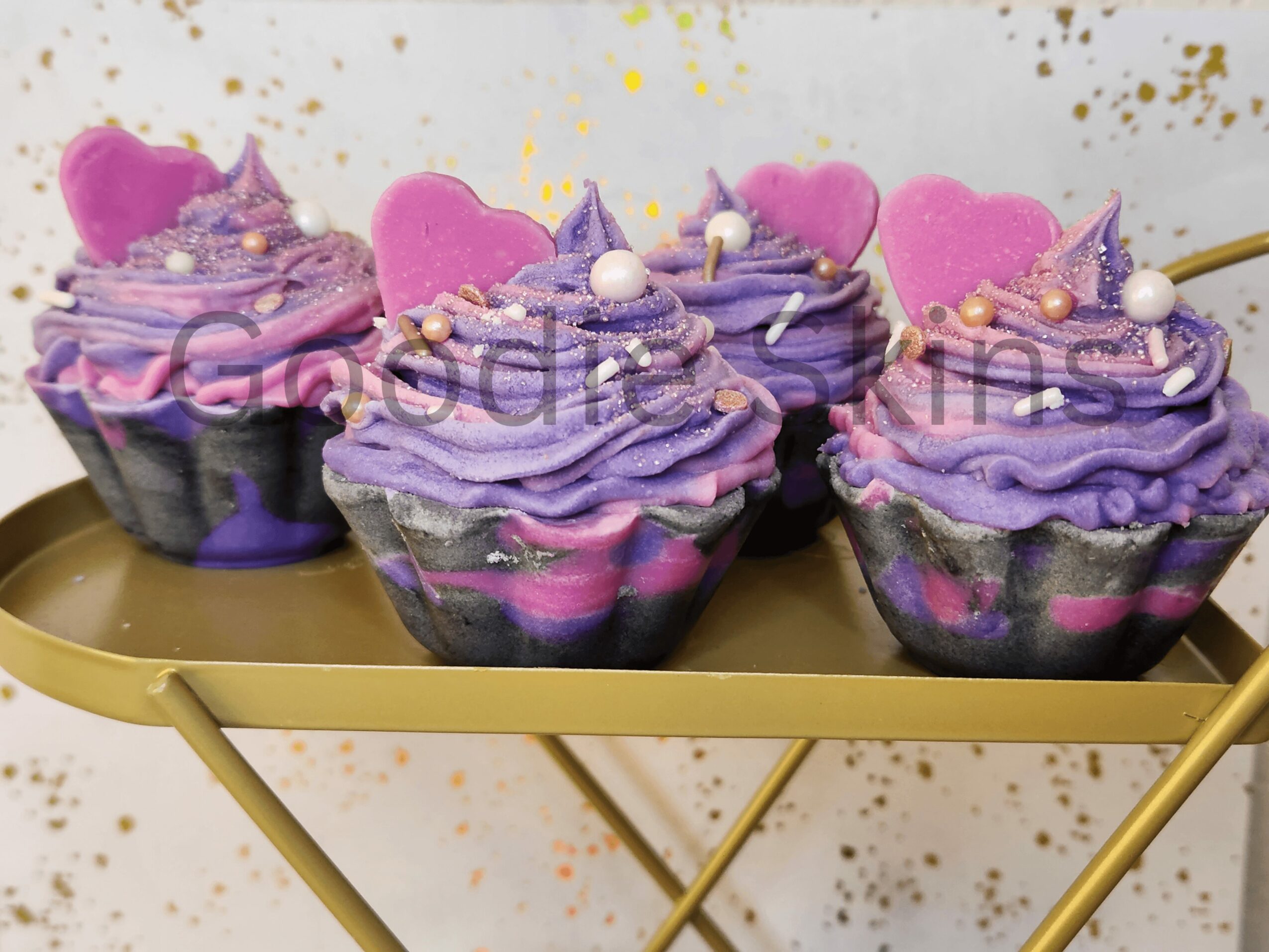 Four purple and pink decorated cupcakes on a gold tray, with a white background and gold splatters.