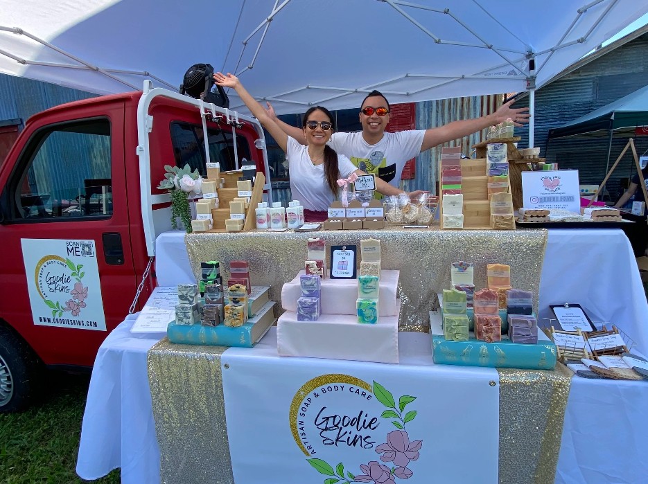 Two people stand behind a table with skincare products at an outdoor market stall, under a canopy, with a red vehicle nearby.