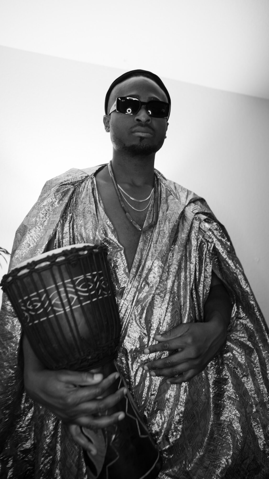 Person wearing sunglasses and traditional clothing, holding a drum, standing against a plain background.