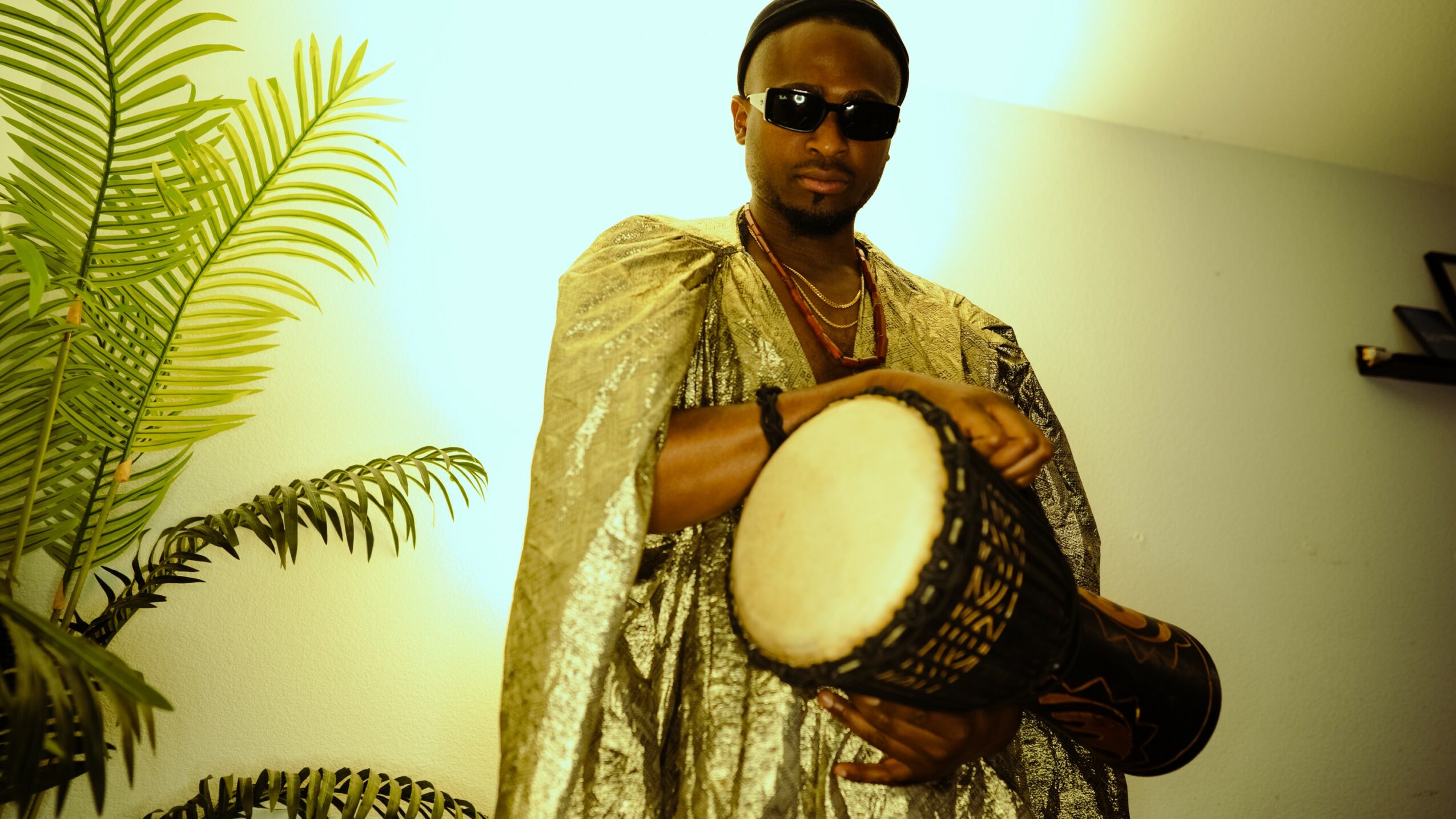 Person wearing sunglasses holding a drum, standing indoors near a plant, with a plain wall background.