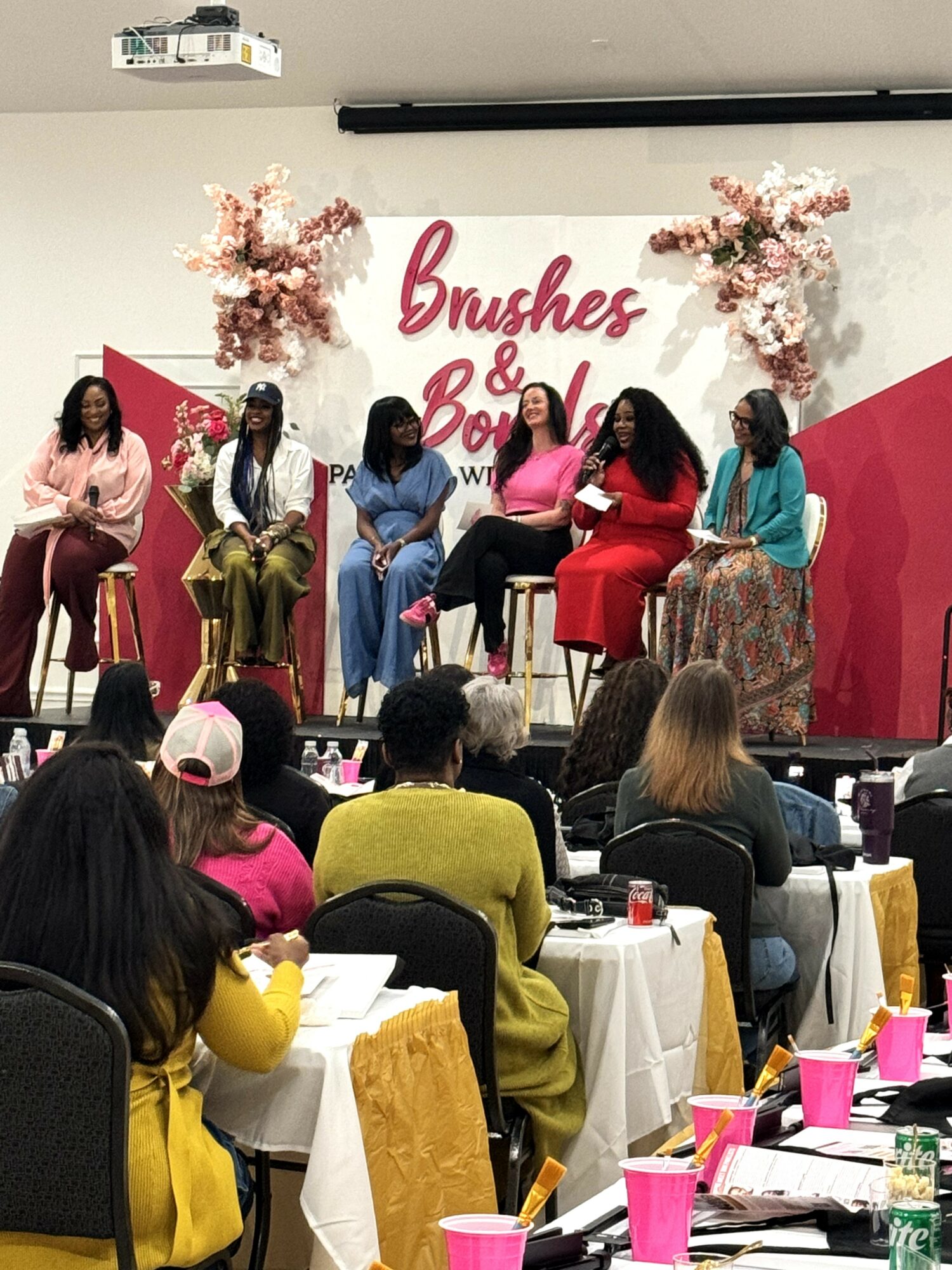 Six women seated on stage during a panel discussion, with a pink and white backdrop and floral decorations.