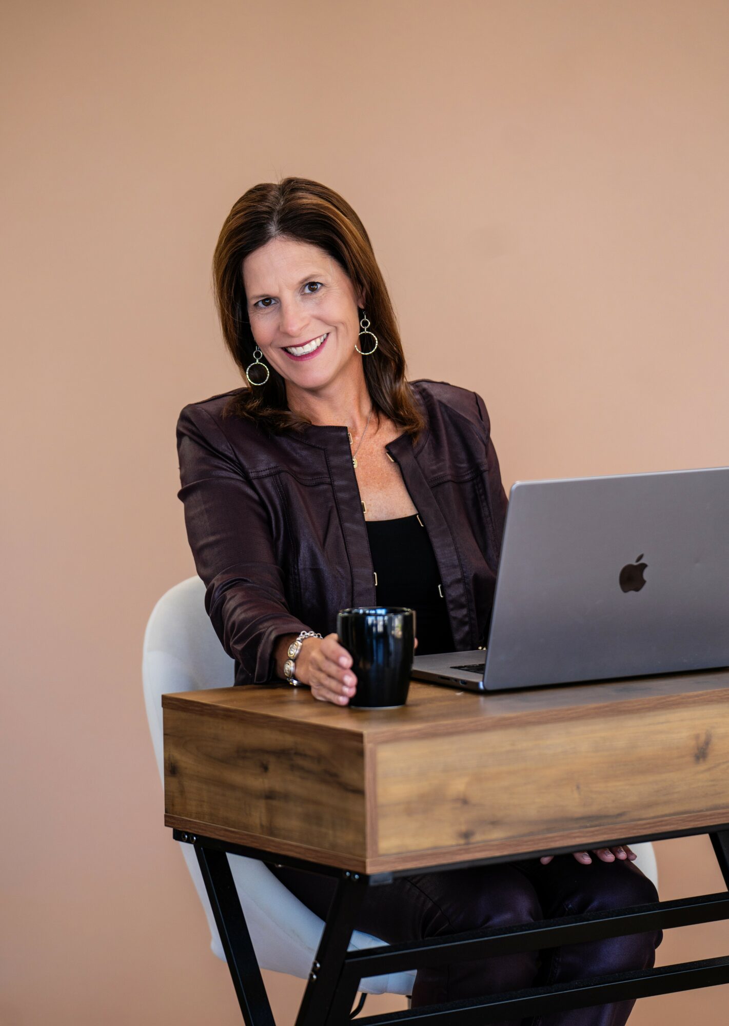 Woman with shoulder-length brown hair, wearing a dark jacket, sitting at a wooden desk with a laptop and a mug.