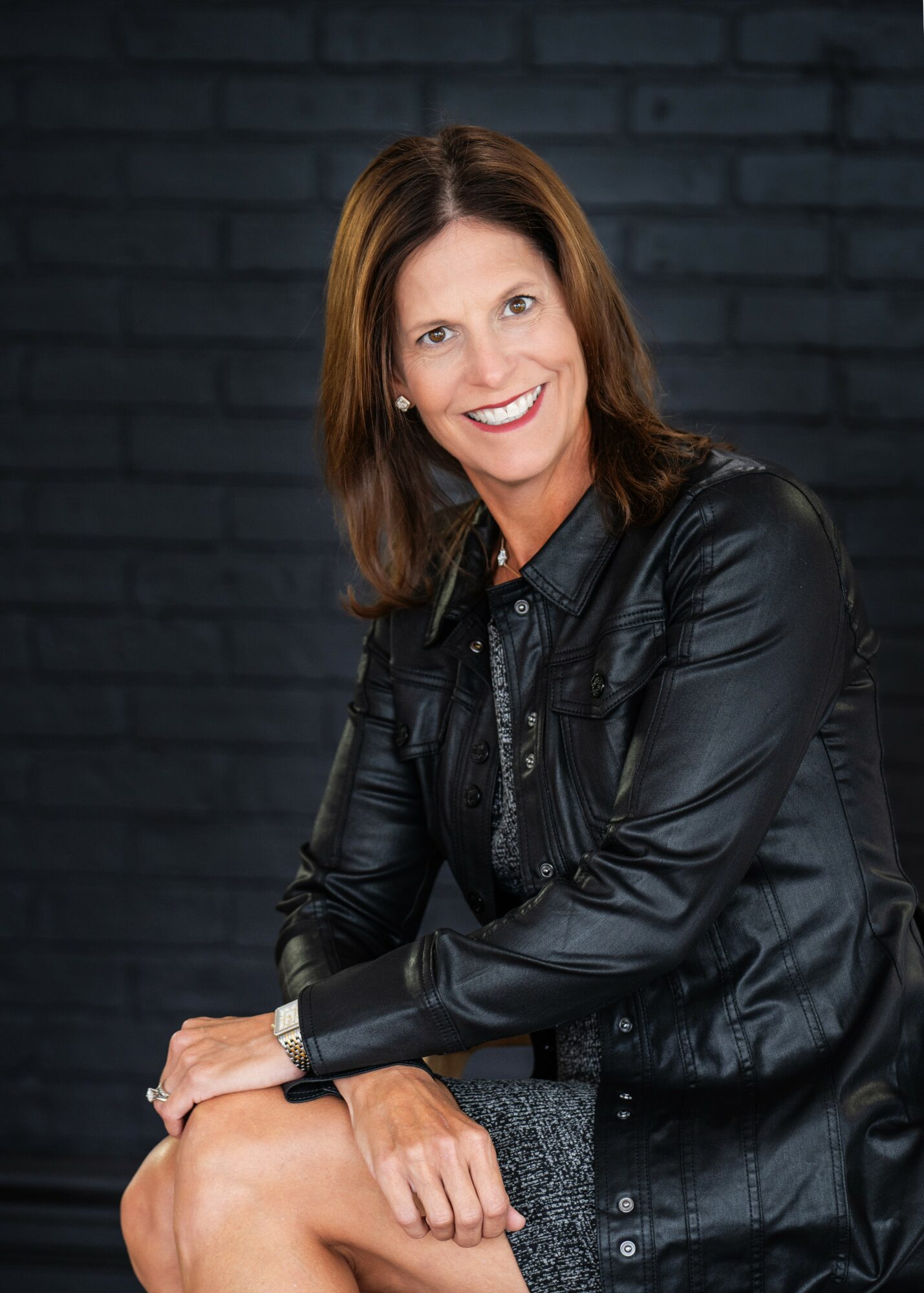 Woman with shoulder-length brown hair smiling, wearing a black leather jacket, sitting against a dark brick wall.