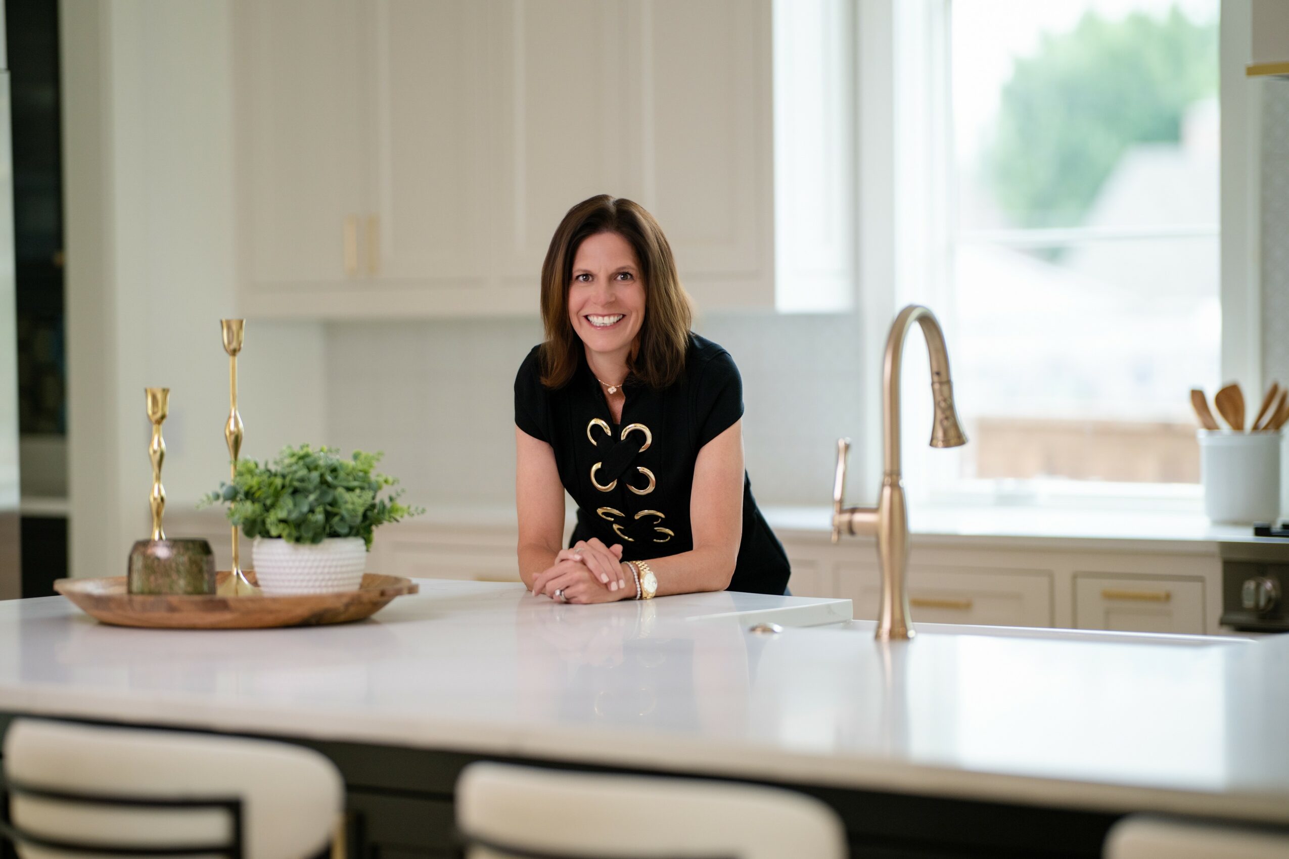 Woman with shoulder-length hair smiling at kitchen counter, with a window and white cabinets in background.