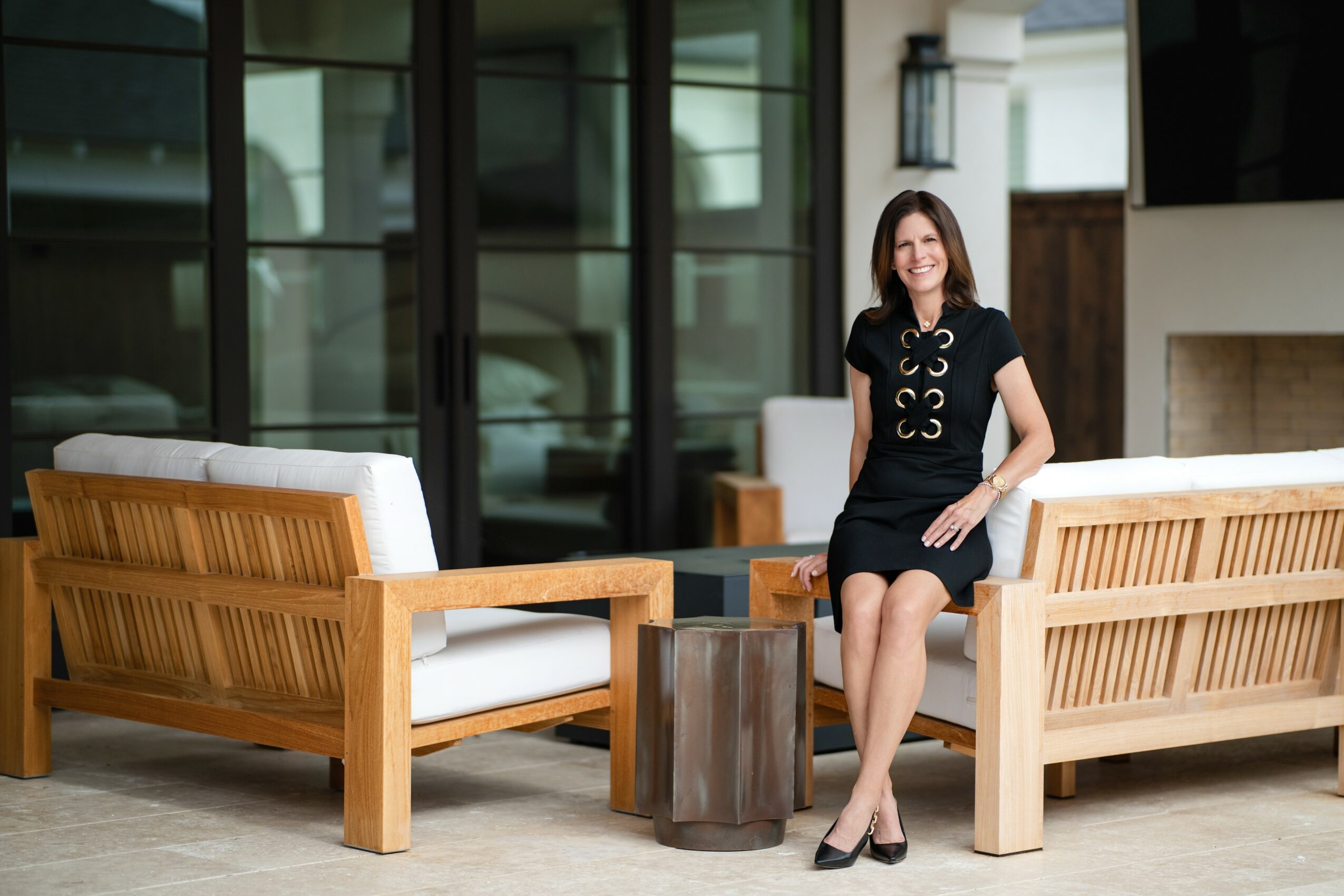 Woman sitting on a wooden outdoor sofa with cushions, smiling, in a patio area with glass windows and a fireplace.