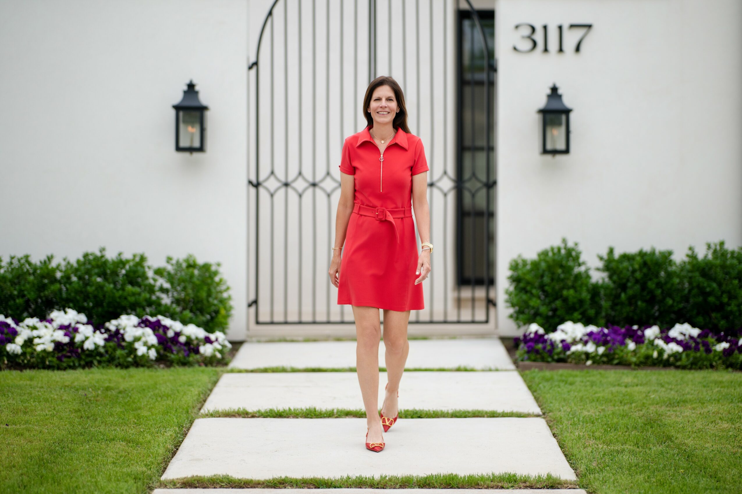 Woman in red dress standing on a pathway in front of a white house with black lanterns and garden flowers.