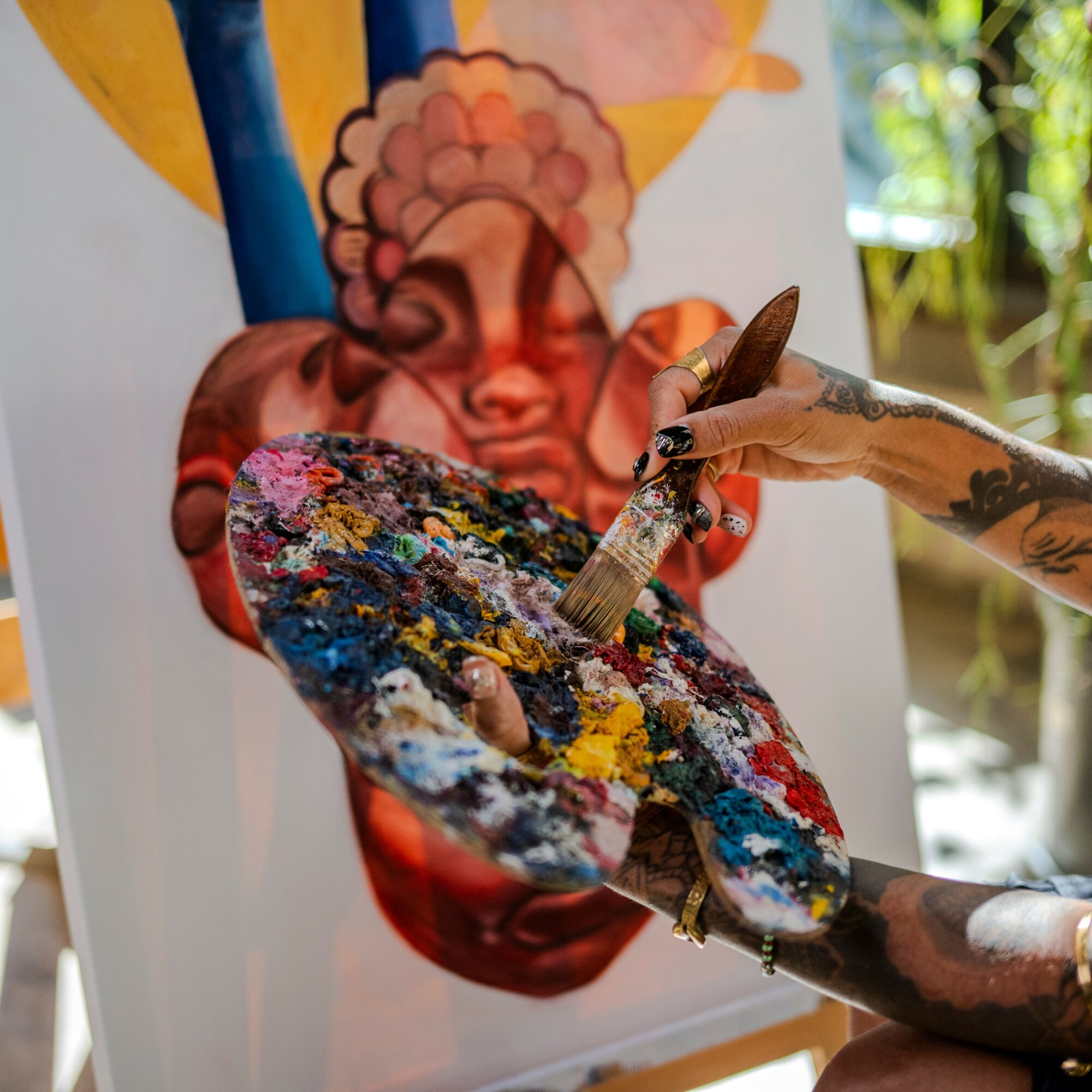 Person with tattooed arm holding a paintbrush and palette, painting a colorful abstract face on a canvas.
