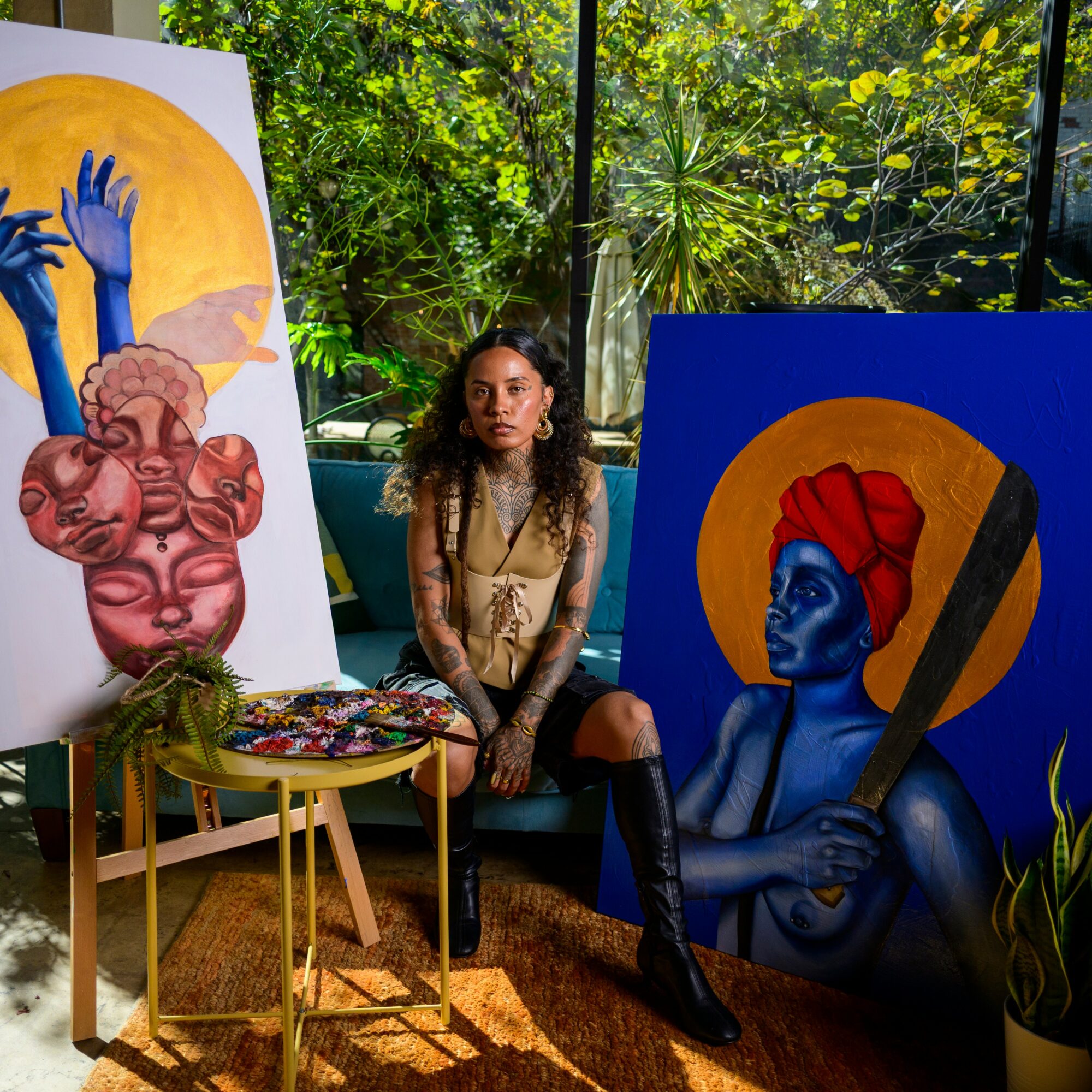 Woman sitting between two colorful paintings of abstract and portrait styles, surrounded by plants and sunlight.