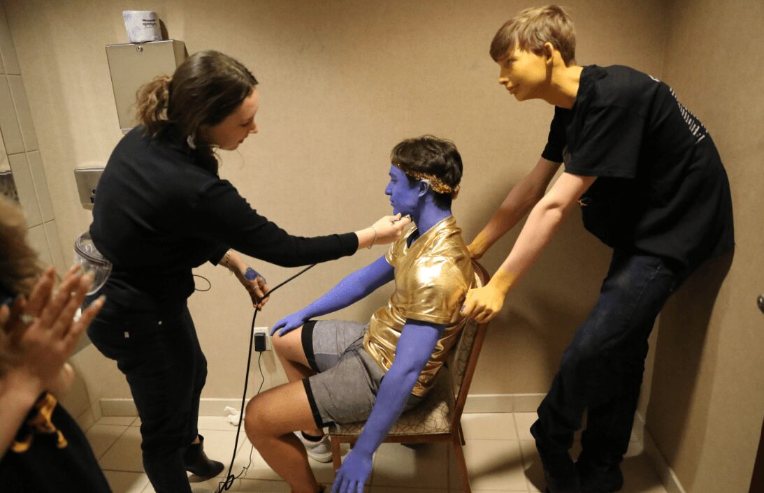 Person with painted blue face and gold shirt seated, two people assisting, one clapping, in a room with beige walls.