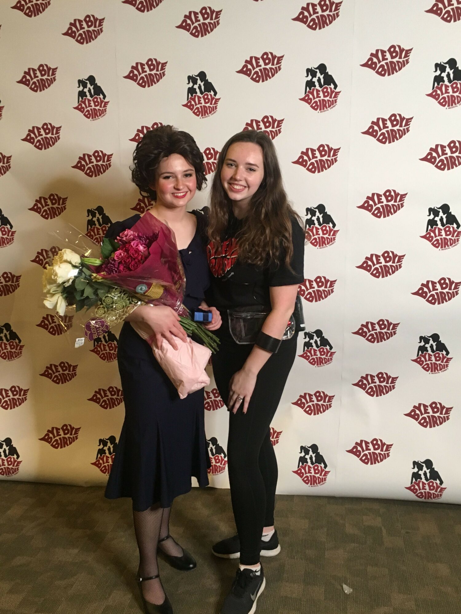 Two women standing in front of a backdrop with a repeating logo pattern, one holding a bouquet of flowers.