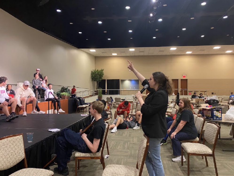Woman standing and pointing at ceiling in a large room with seated people and a stage, some people wearing pink, some in red, and others in casual clothing.