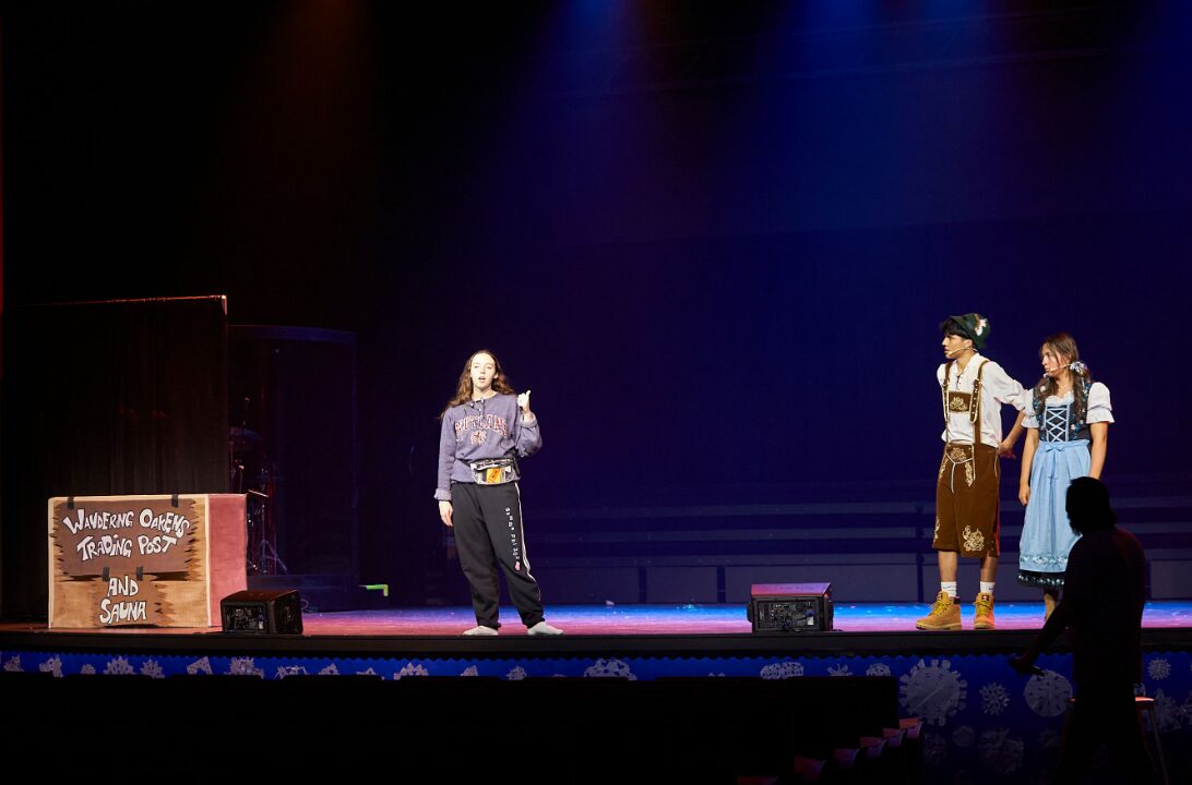 Three people on stage, one speaking, two listening, with a sign on the left and a dark background.
