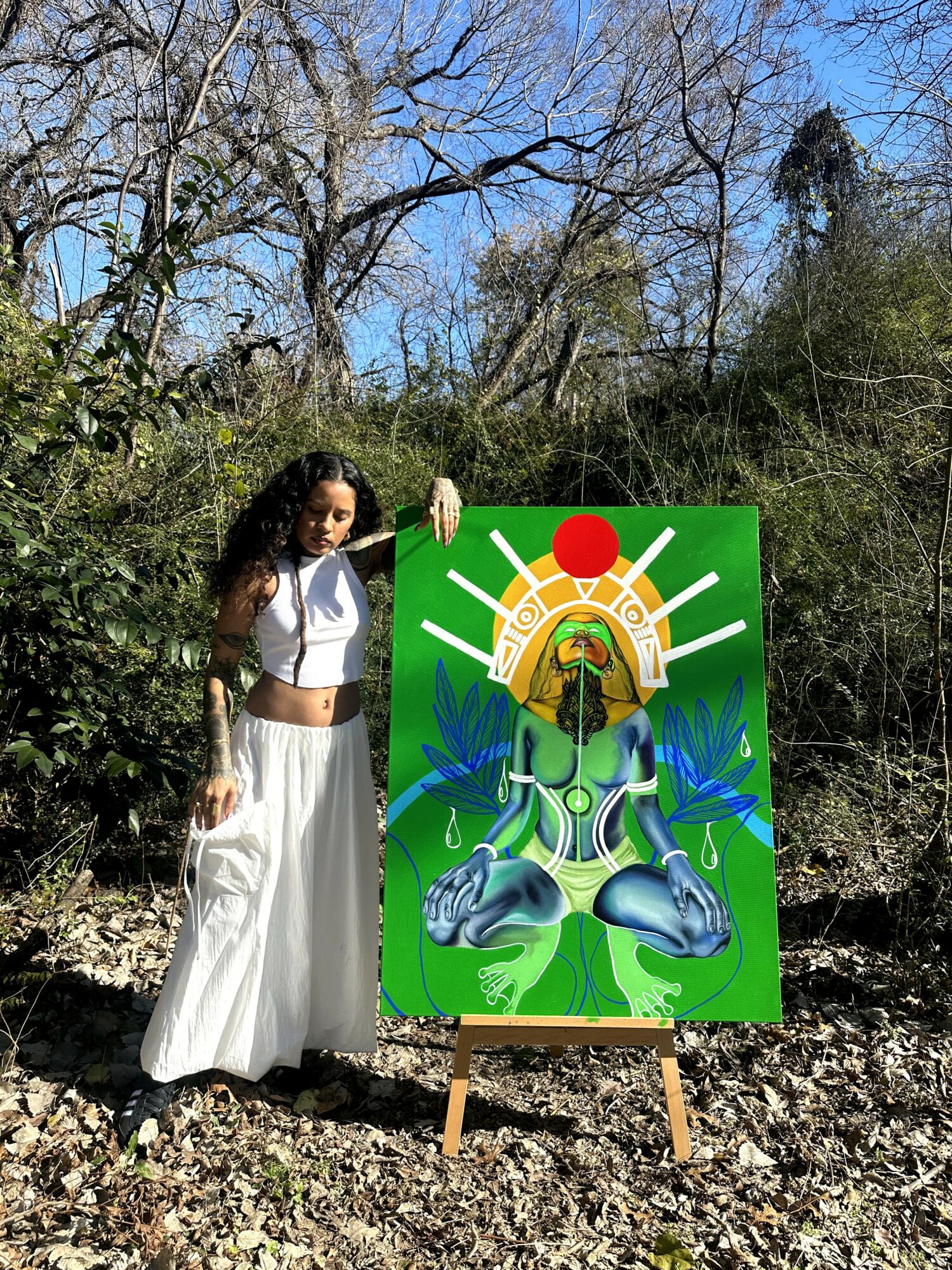 Woman with curly hair in white outfit standing outdoors next to a colorful painting of a meditative figure with a red circle above head, surrounded by blue leaves, on a green background.
