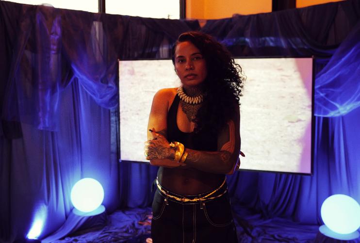 A woman with curly hair stands in front of a large screen, with blue lighting and round lights on stage.