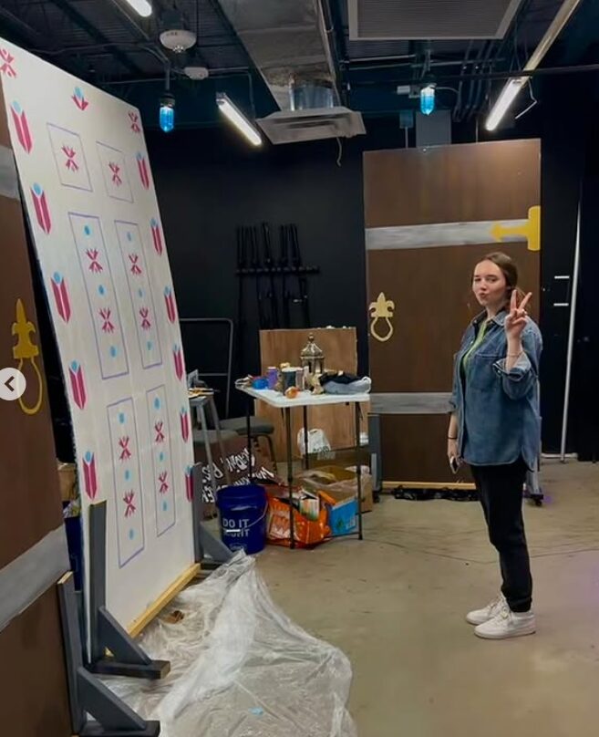 Person taking a selfie in a room with black walls, a whiteboard, and various items on a table, making a peace sign.