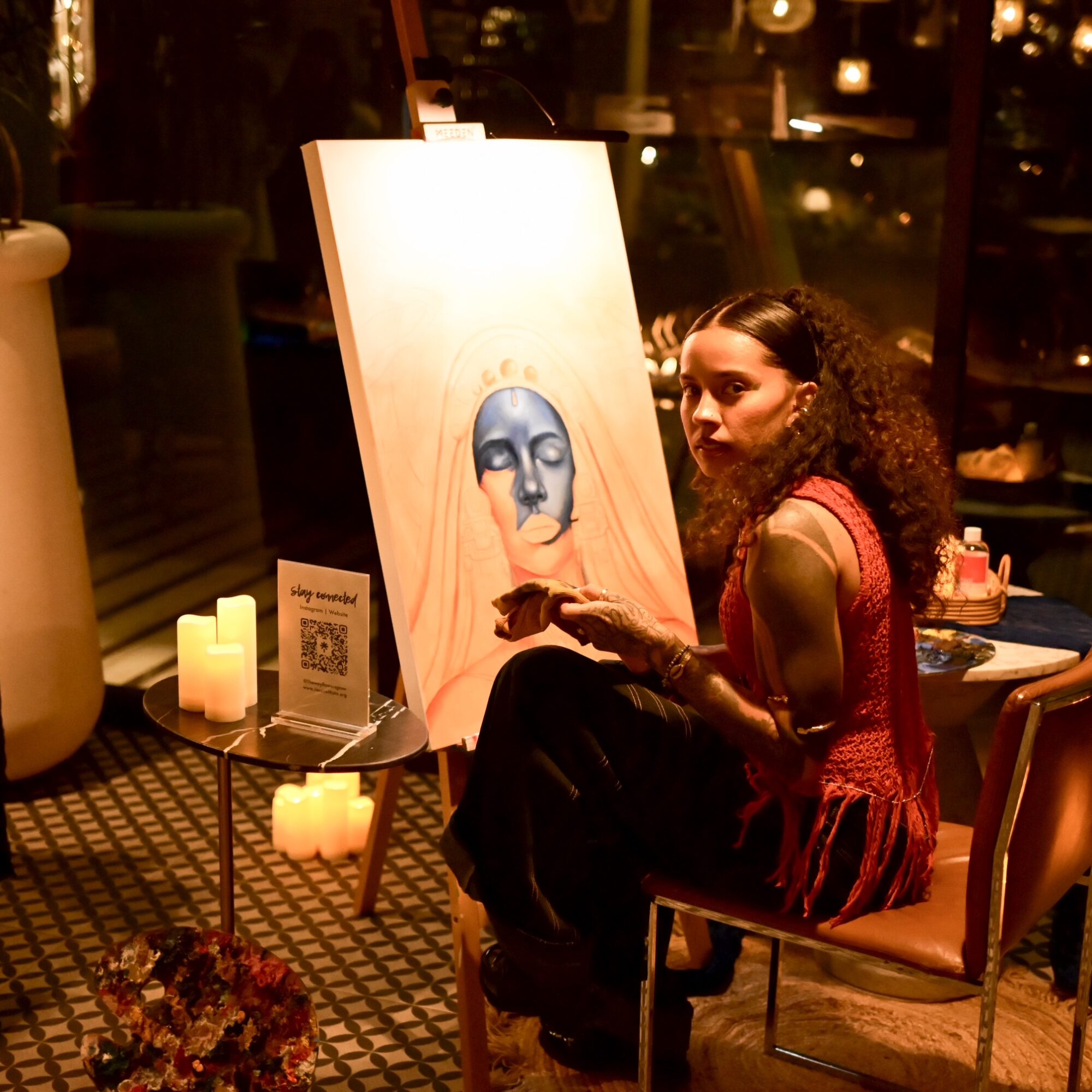 Woman with curly hair sitting next to a painting of a face with blue makeup, in a warmly lit room with candles.