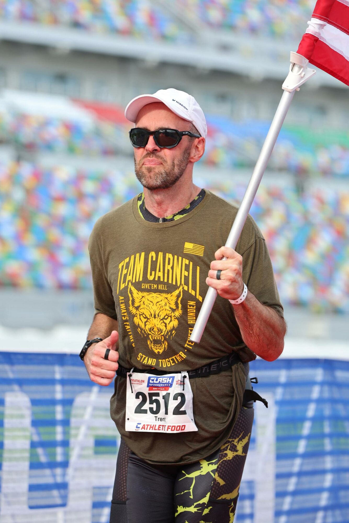 Man wearing sunglasses, a cap, and a T-shirt holding a flag at a race event, with a crowd in the background.