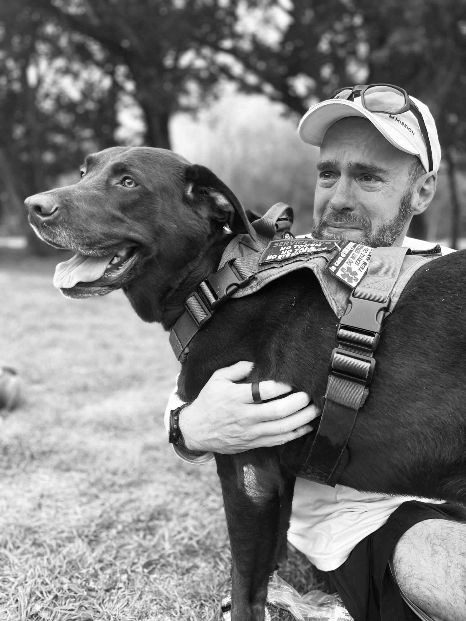 Man holding a dog outdoors, both looking to the side, trees in background, black and white photo.