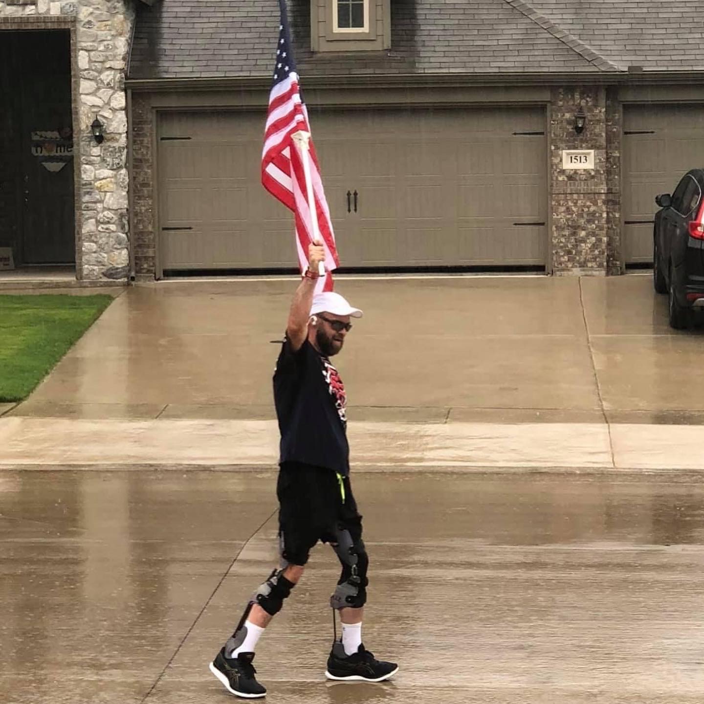 Child walking on wet driveway holding American flag, wearing a cap, black shorts, and sneakers, with a house and garage in background.