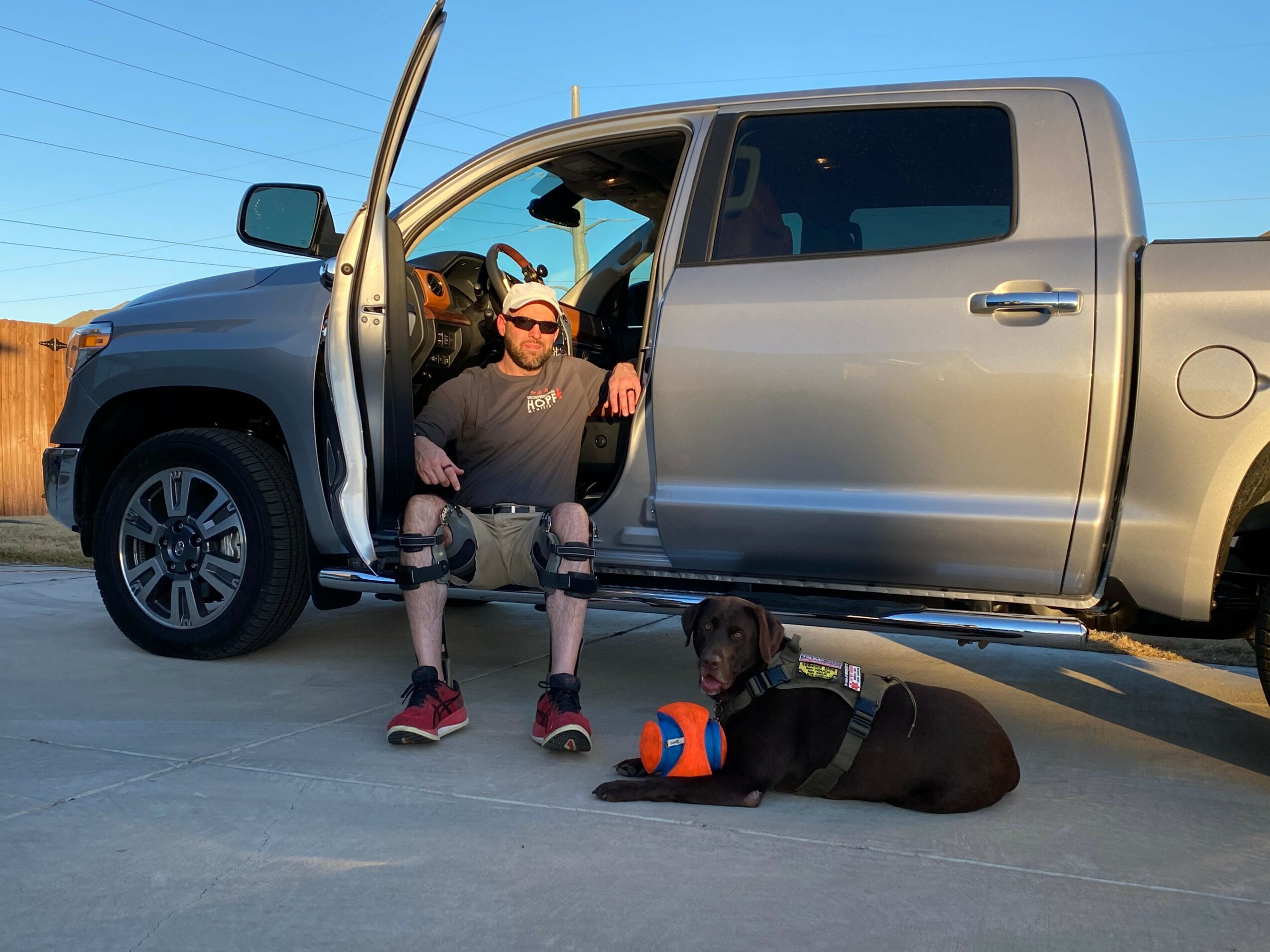 Man sitting on the ground with a dog lying nearby, vehicle door open behind him, clear blue sky.