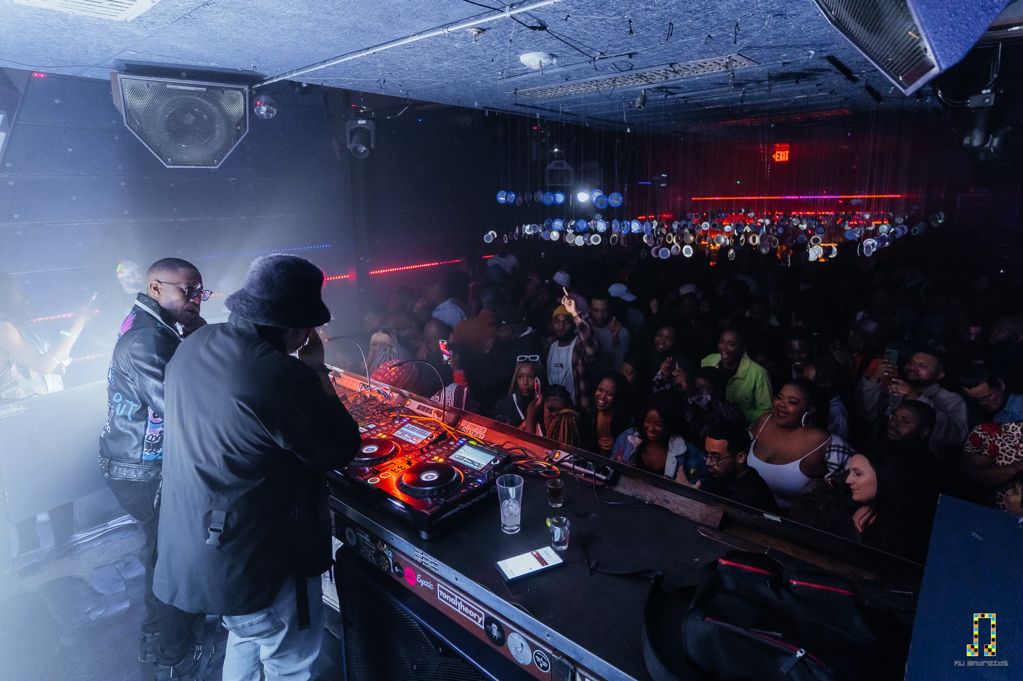 Two DJs perform on stage with a large crowd dancing in a club, colorful lights and fog effects present.