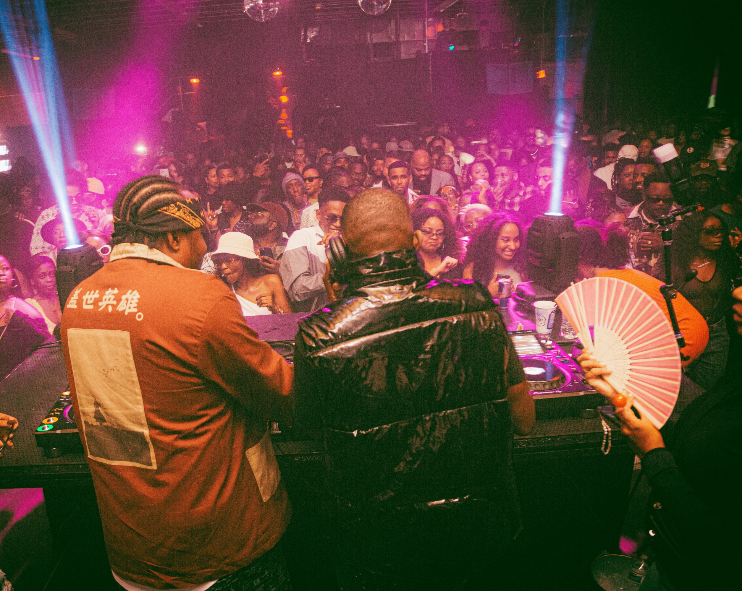 Two DJs perform at a nightclub with a crowd dancing under colorful lights, some holding fans, in a lively scene.