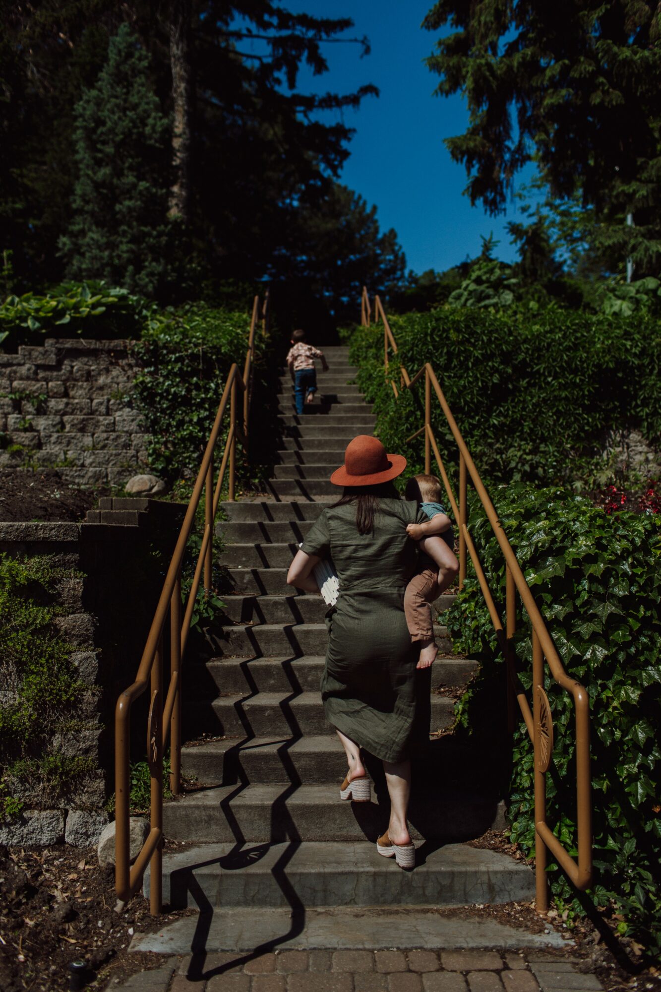 People walking up outdoor stone stairs with metal railings, surrounded by greenery and trees, under a blue sky.