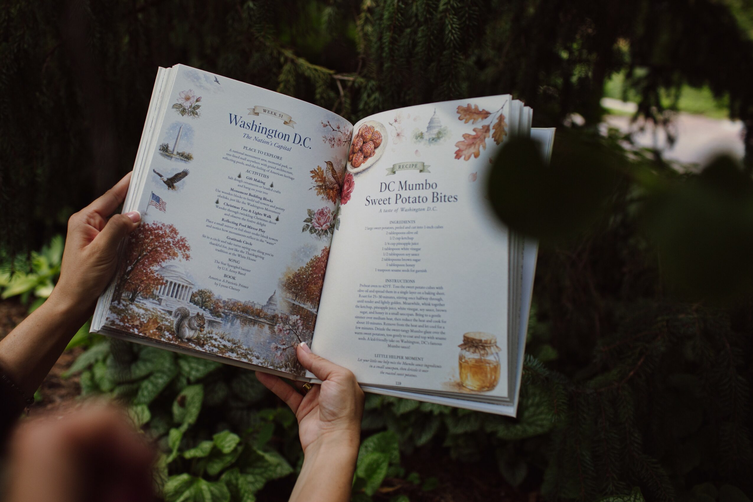 Person holding open cookbook outdoors with floral and food illustrations, background of trees and greenery.